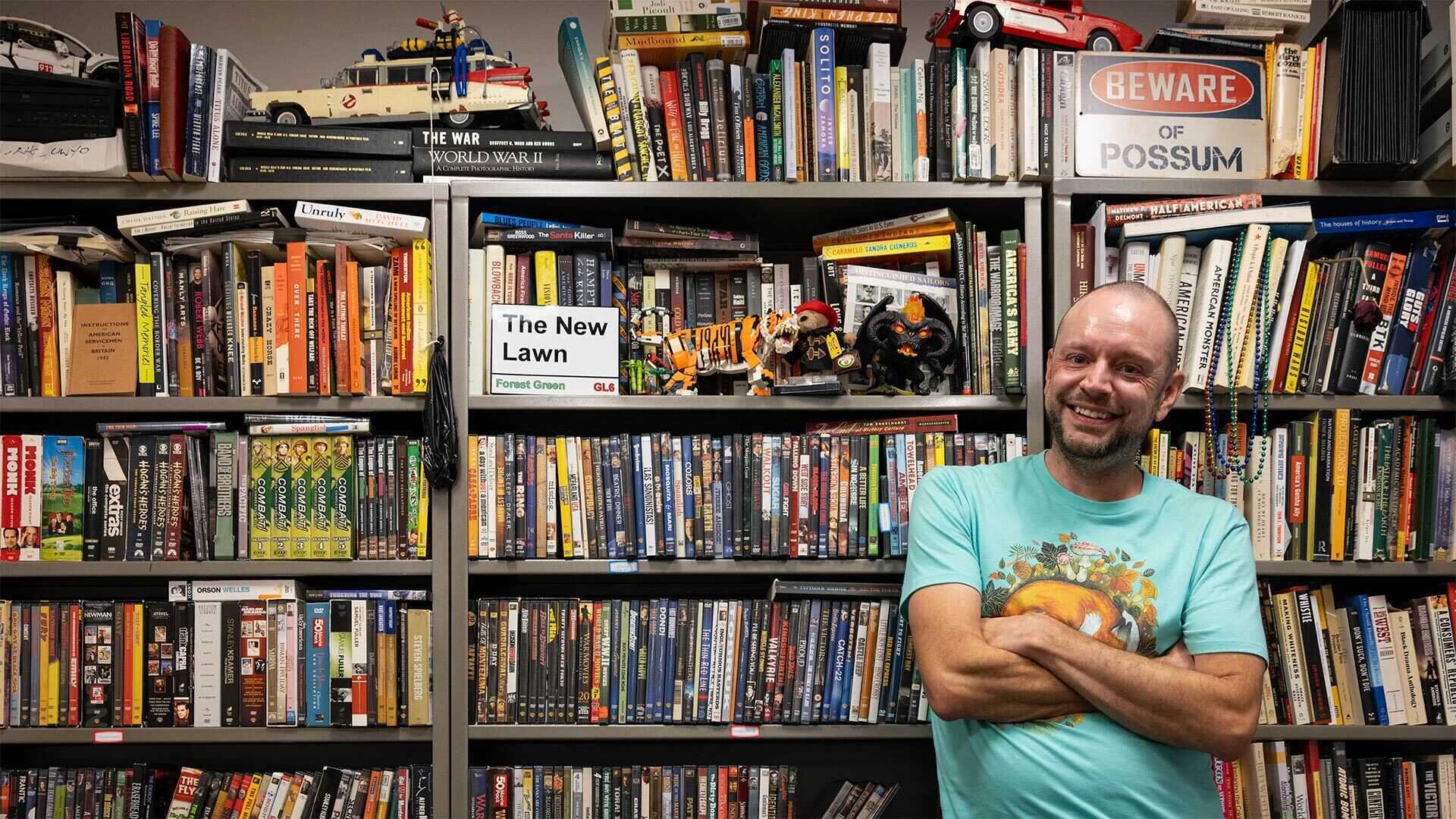 Robert Chester stands in front of bookshelves filled with DVDs and books, wearing a teal shirt and crossing his arms