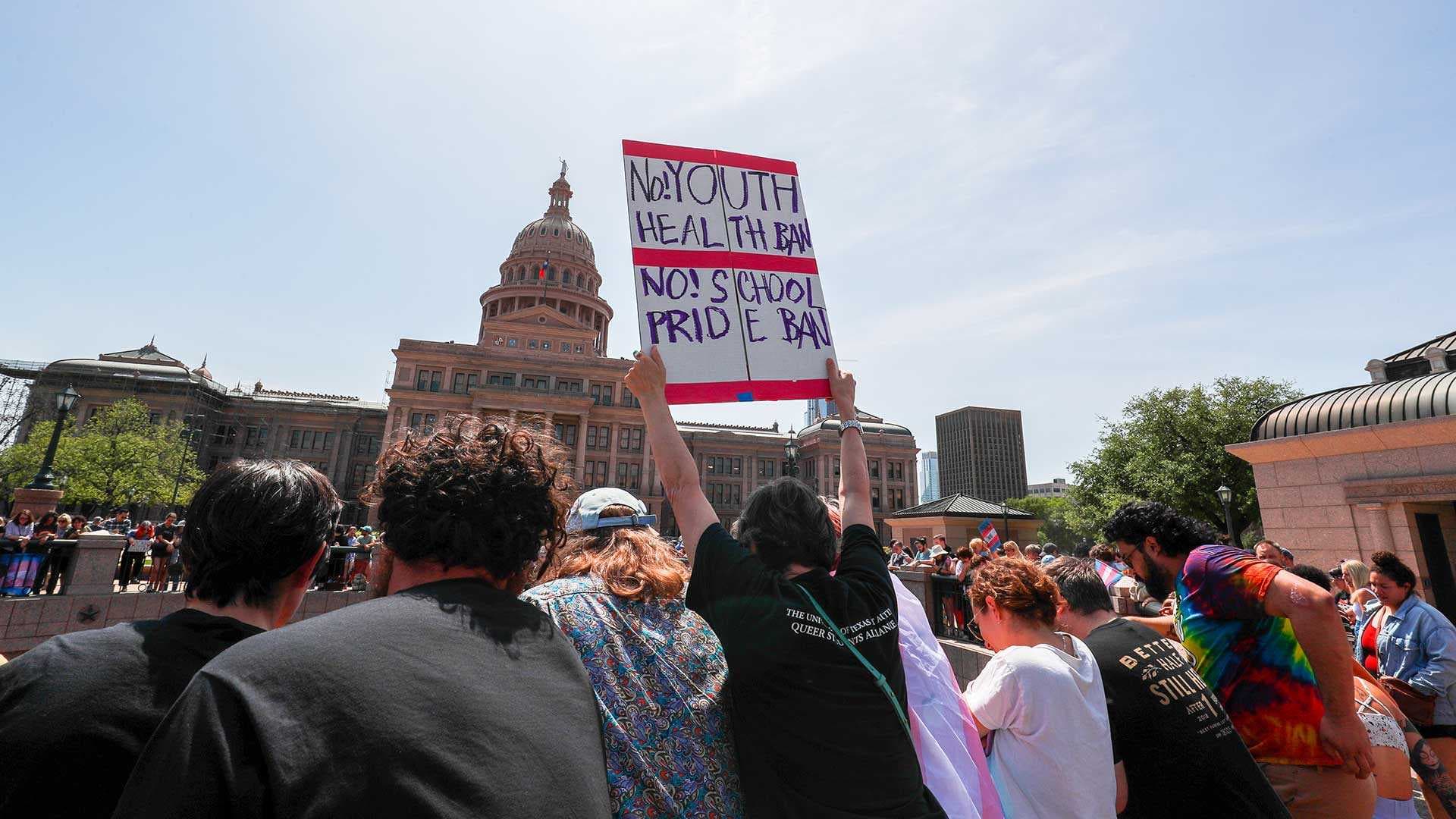 Protesters at the Texas State Capitol building. One holds a sign that reads, "No Youth Health Ban. No School Pride Ban"