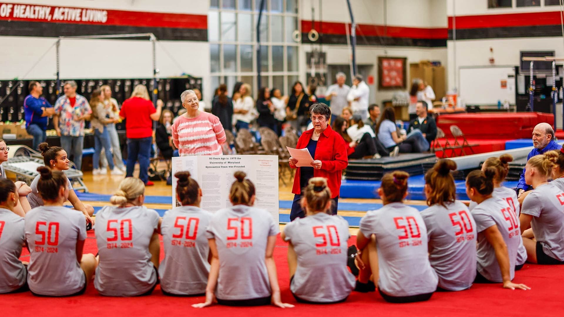Two older women speak to a team of seated younger women in a gym