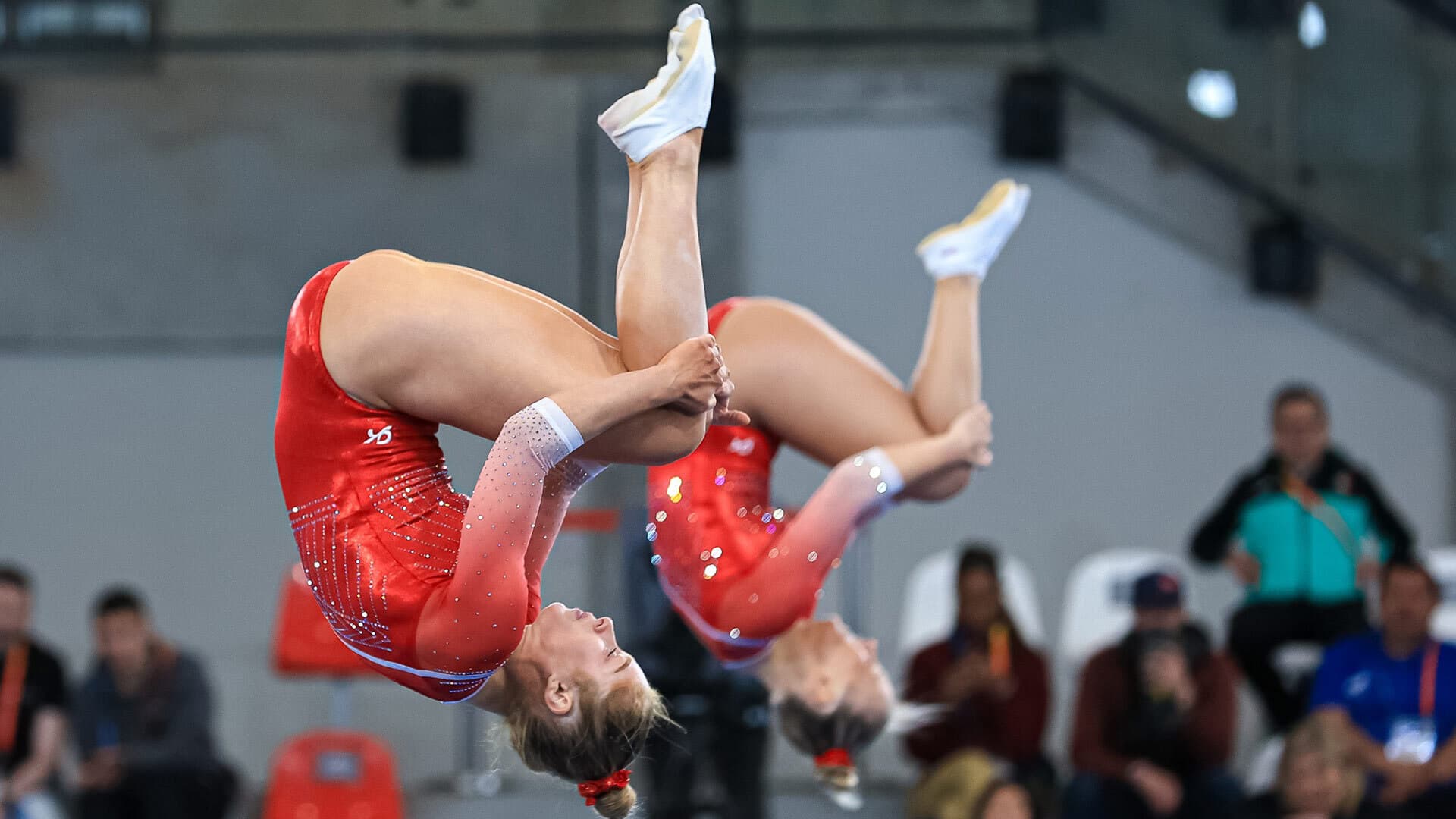 two female athletes flip through the air, upside-down in red leotards