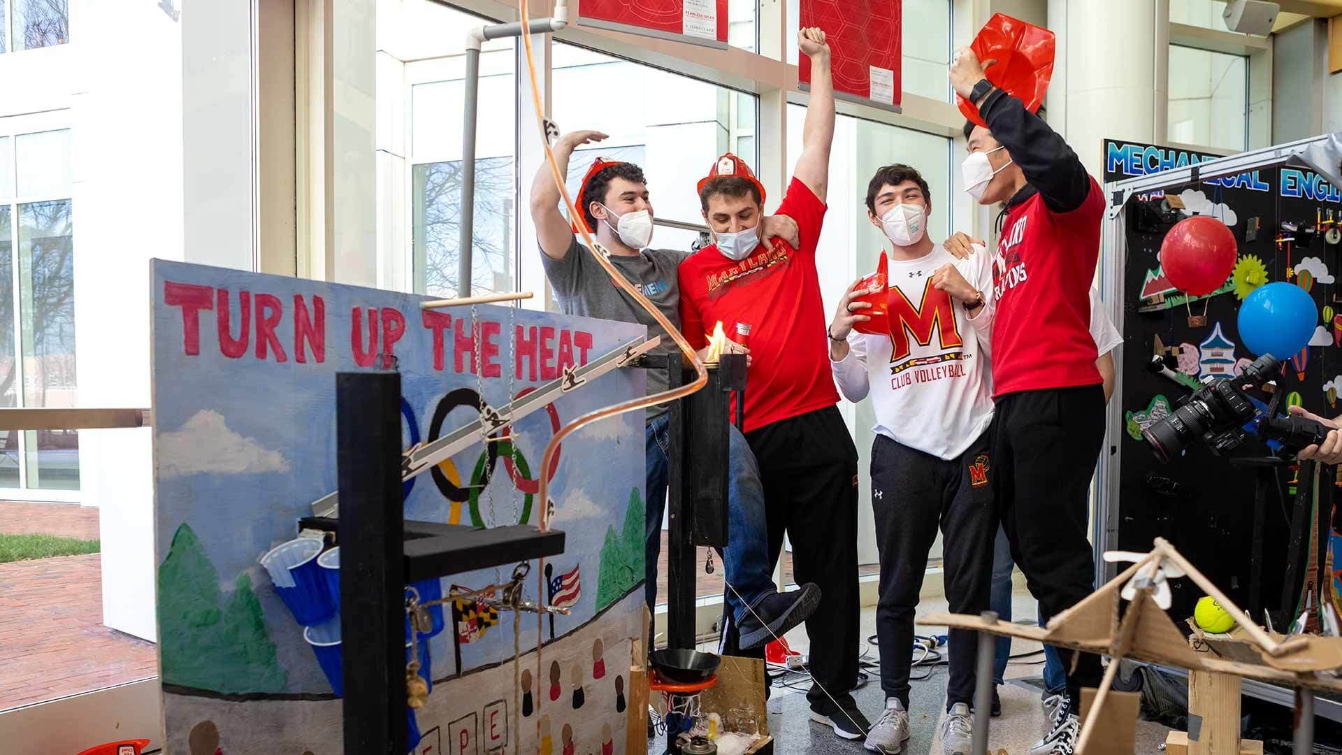 Four students in red firefighter hats celebrate by their Rube Goldberg machine