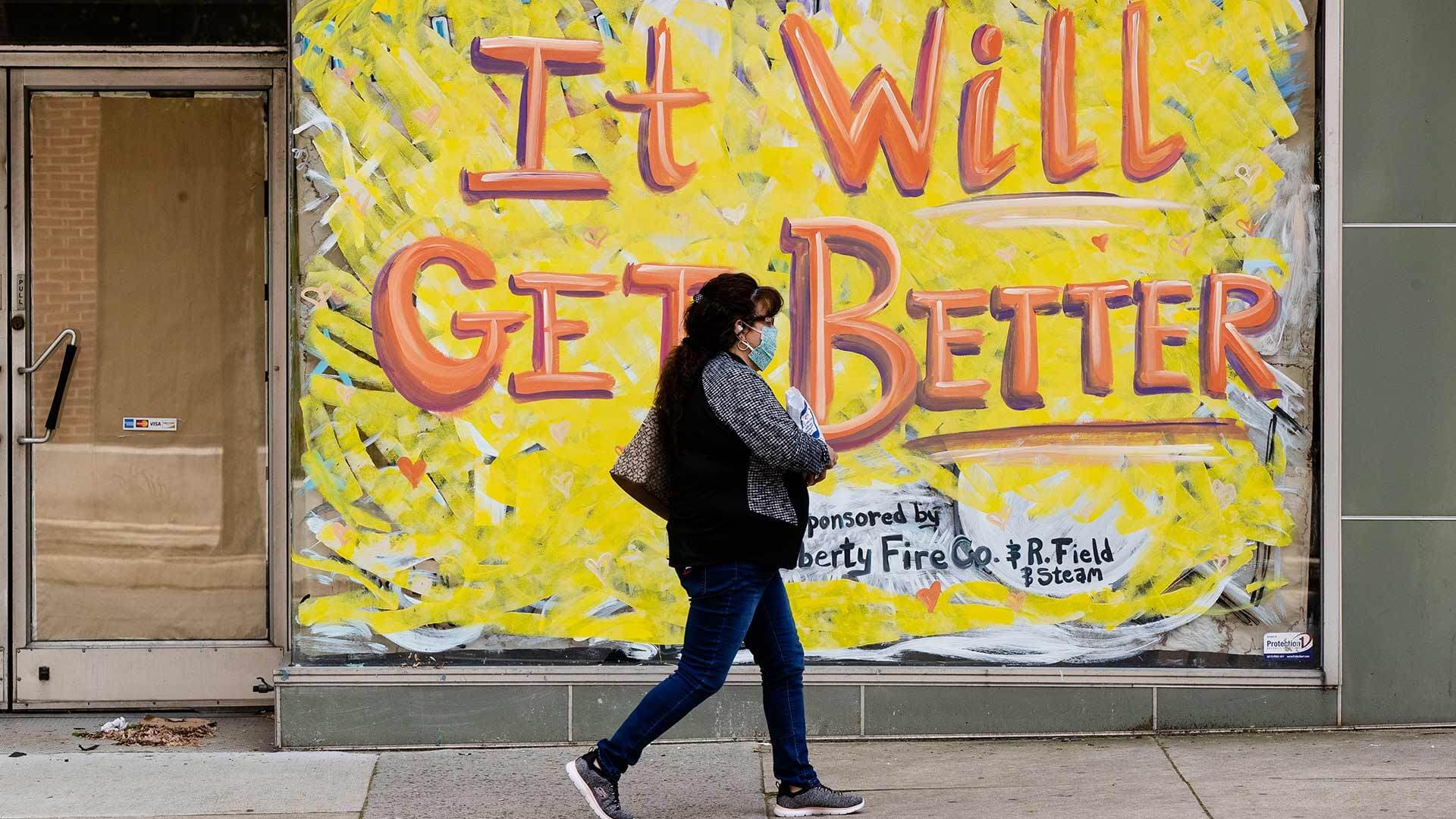Person wearing mask walks by a sign that says "It will get better."
