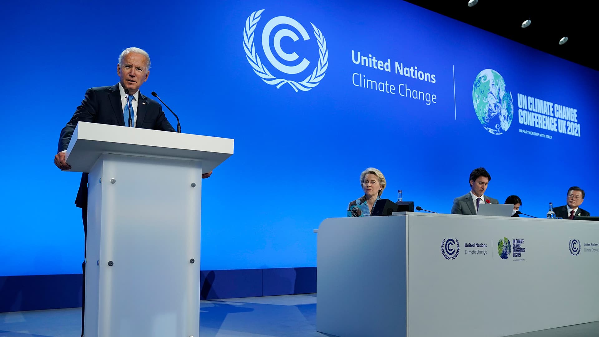 Joe Biden speaking at a podium, with other world leaders seated next to him, in front of a blue screen with the words United Nations Climate Change