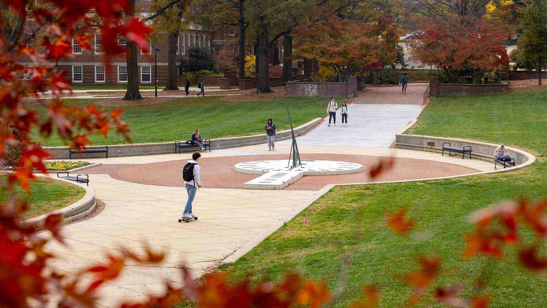 Red leaves frame students walking near the sundial