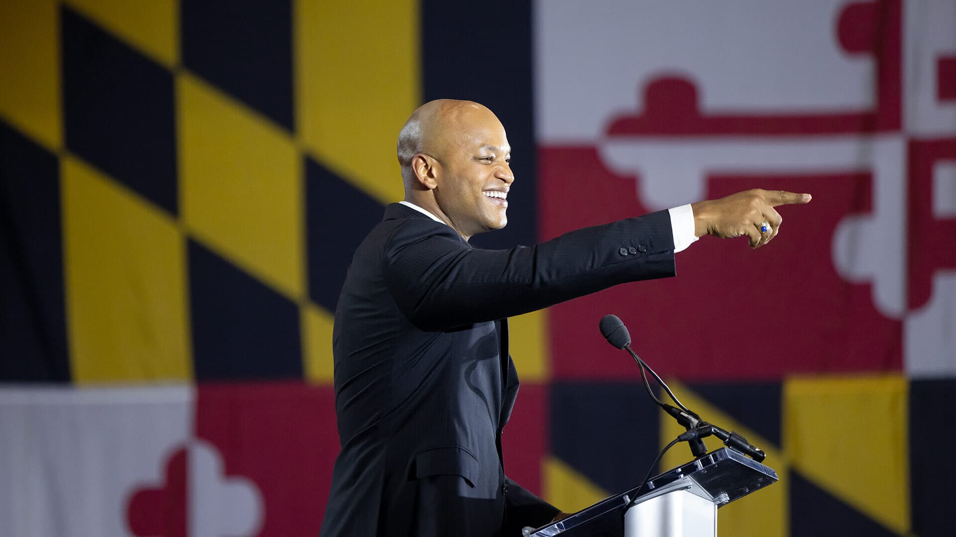 smiling man on stage in front of giant maryland flag points at audience