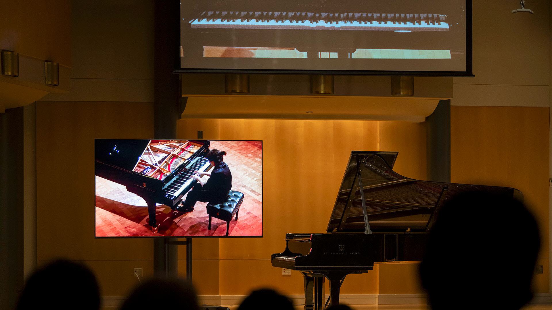 A robot piano plays in a concert hall, while a screen depicts a pianist playing on a piano elsewhere
