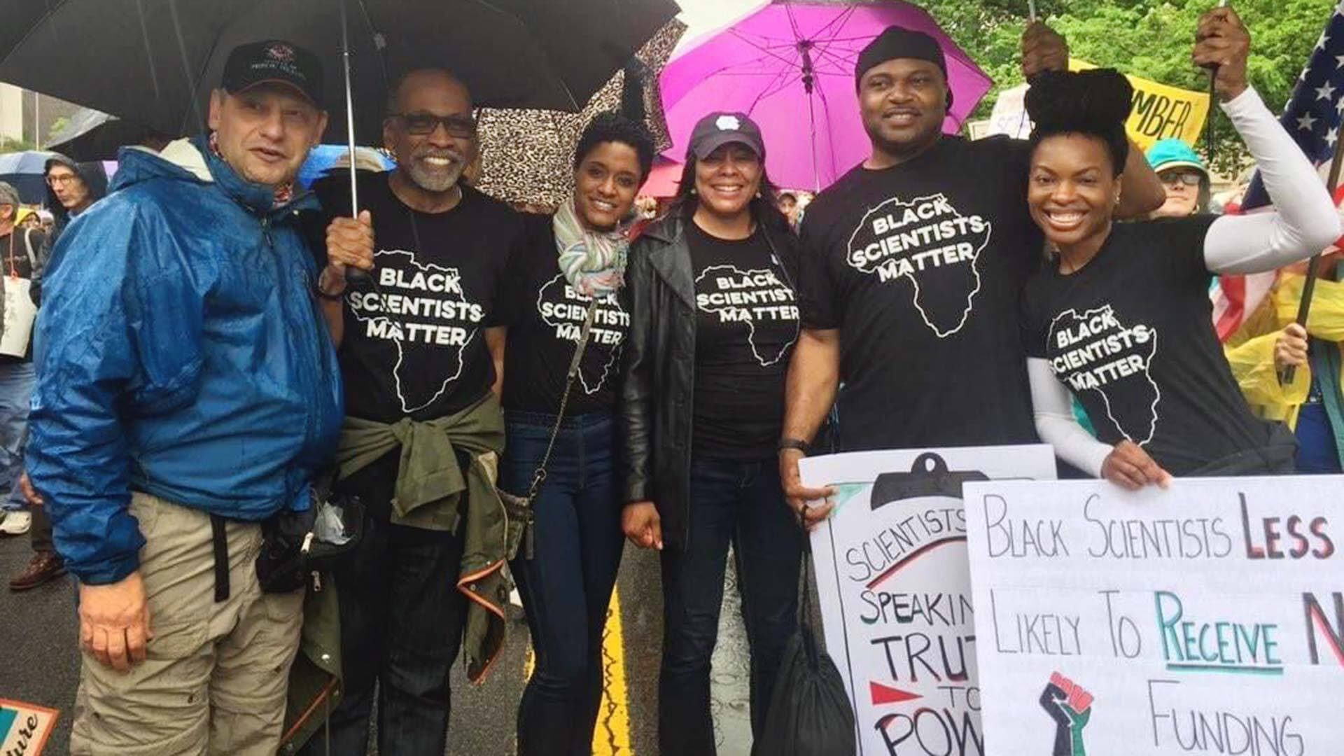 People wearing "Black Scientists Matter" shirts hold umbrellas