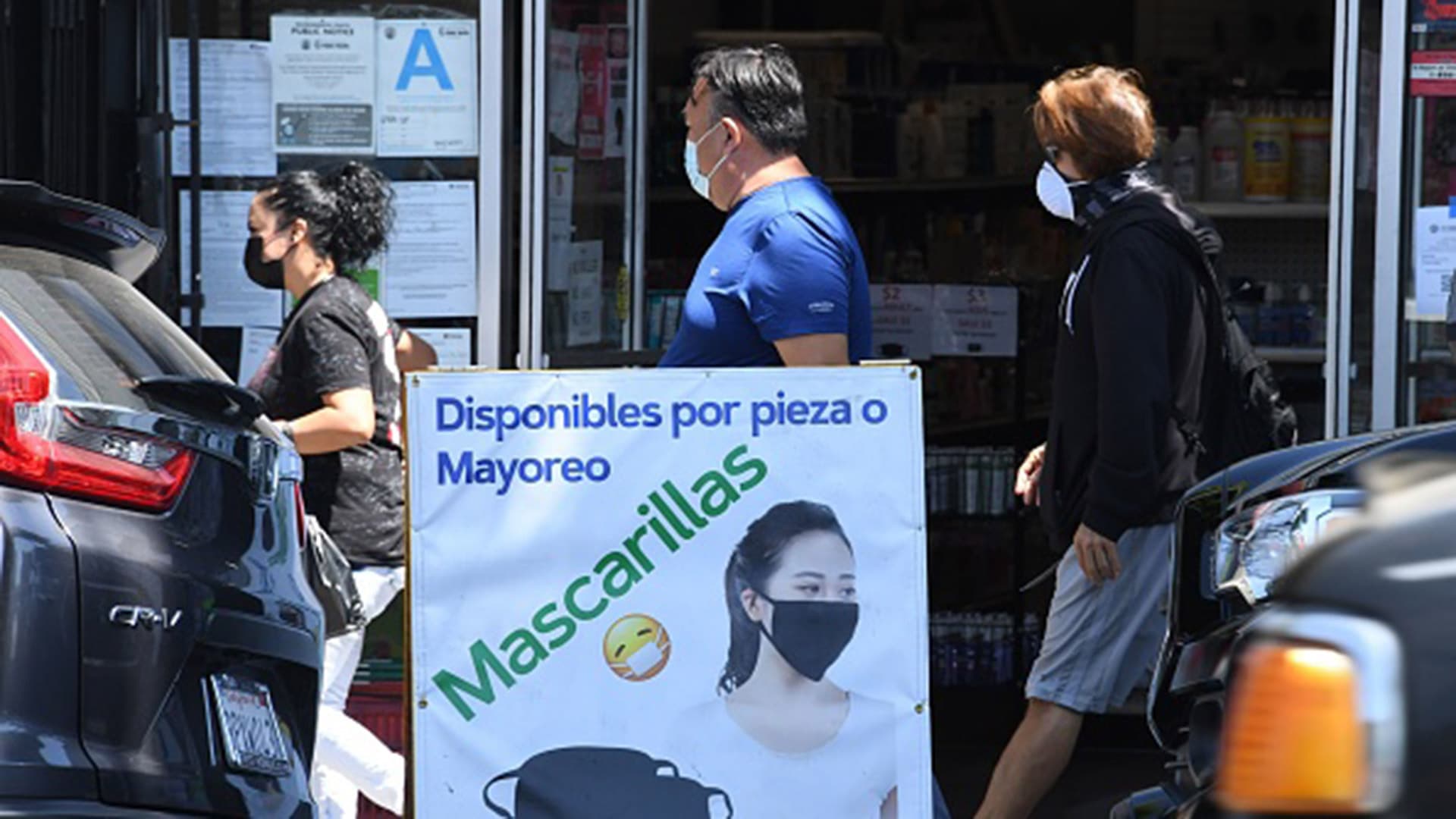 People wearing masks walk by a store with a sign that advertises face coverings in Spanish