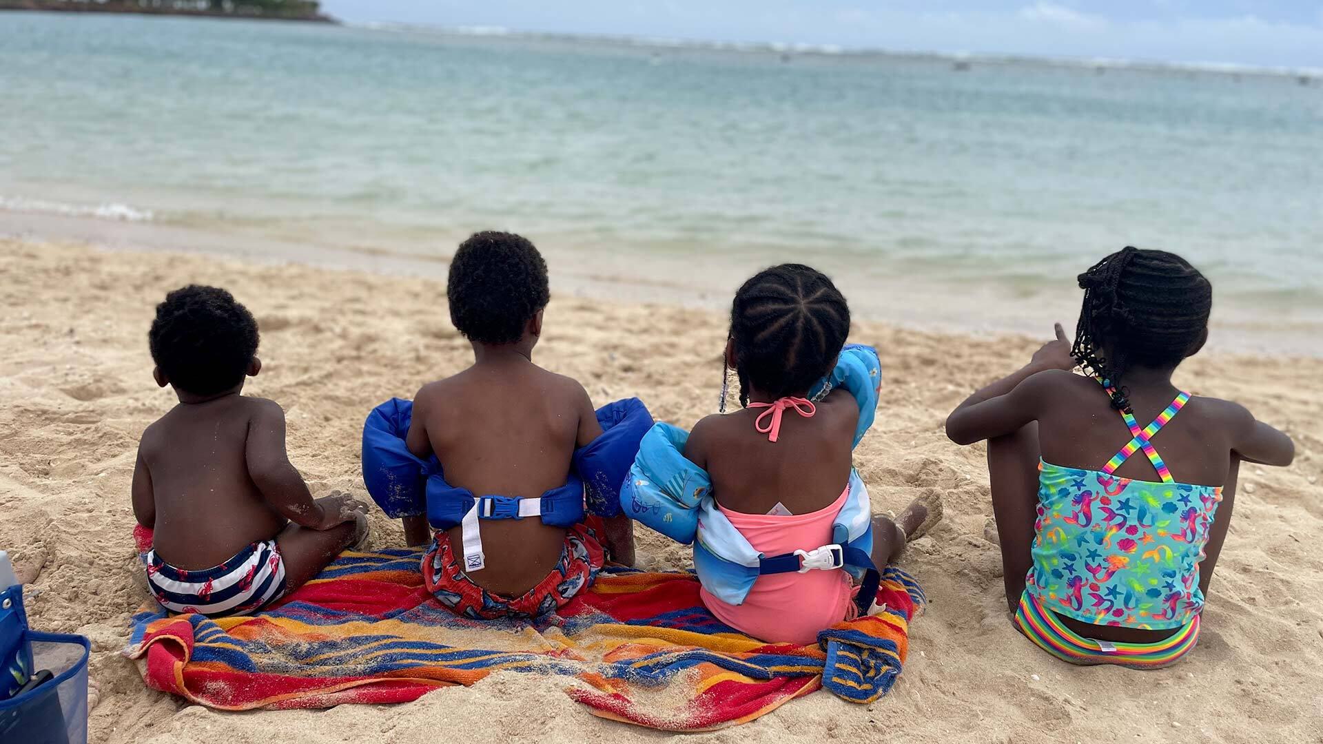 The back of four kids in swimsuits sitting on the beach