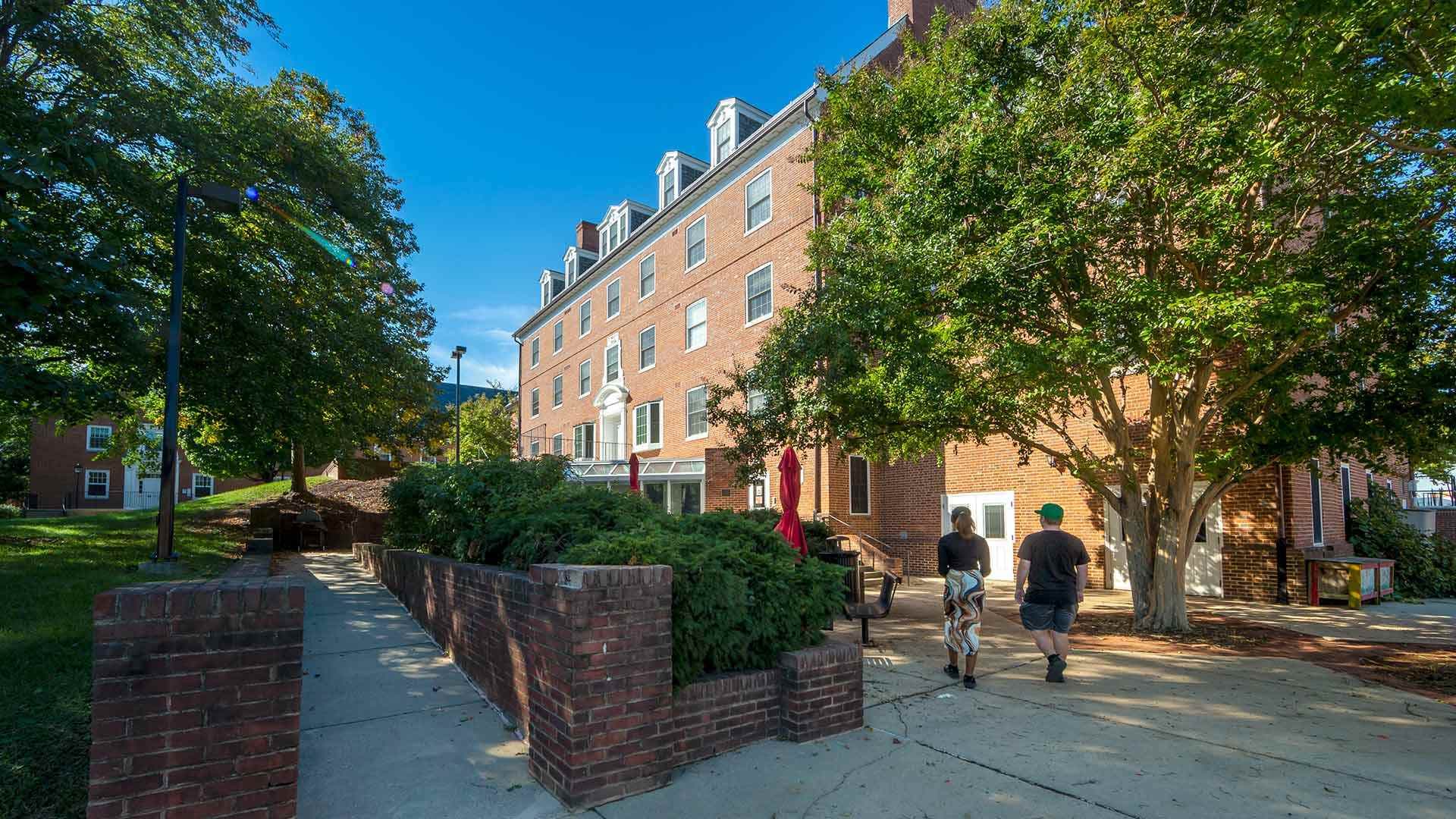 Two students walking toward a brick building surrounded by trees
