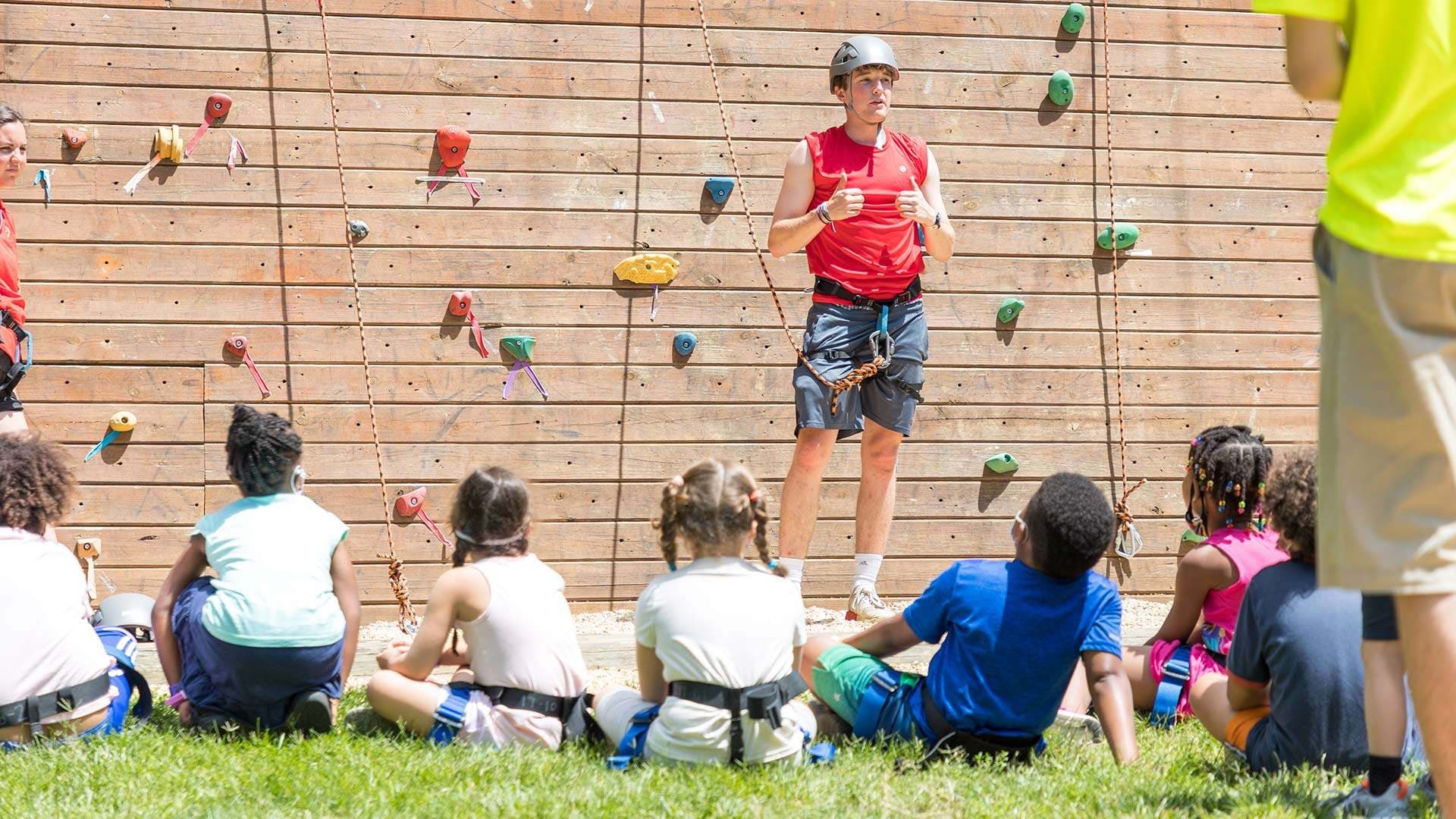 Camp counselor in helmet and gear in front of climbing wall speaking to seated kids