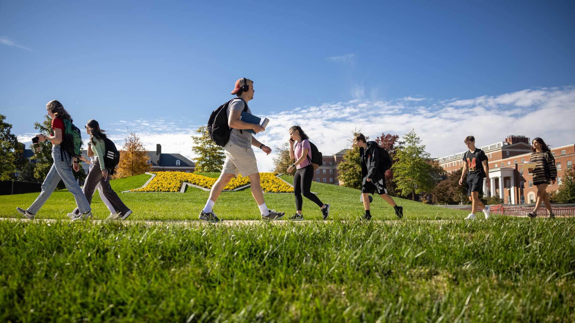 Students walk in front of the a yellow flower-covered M planted on a grass mound