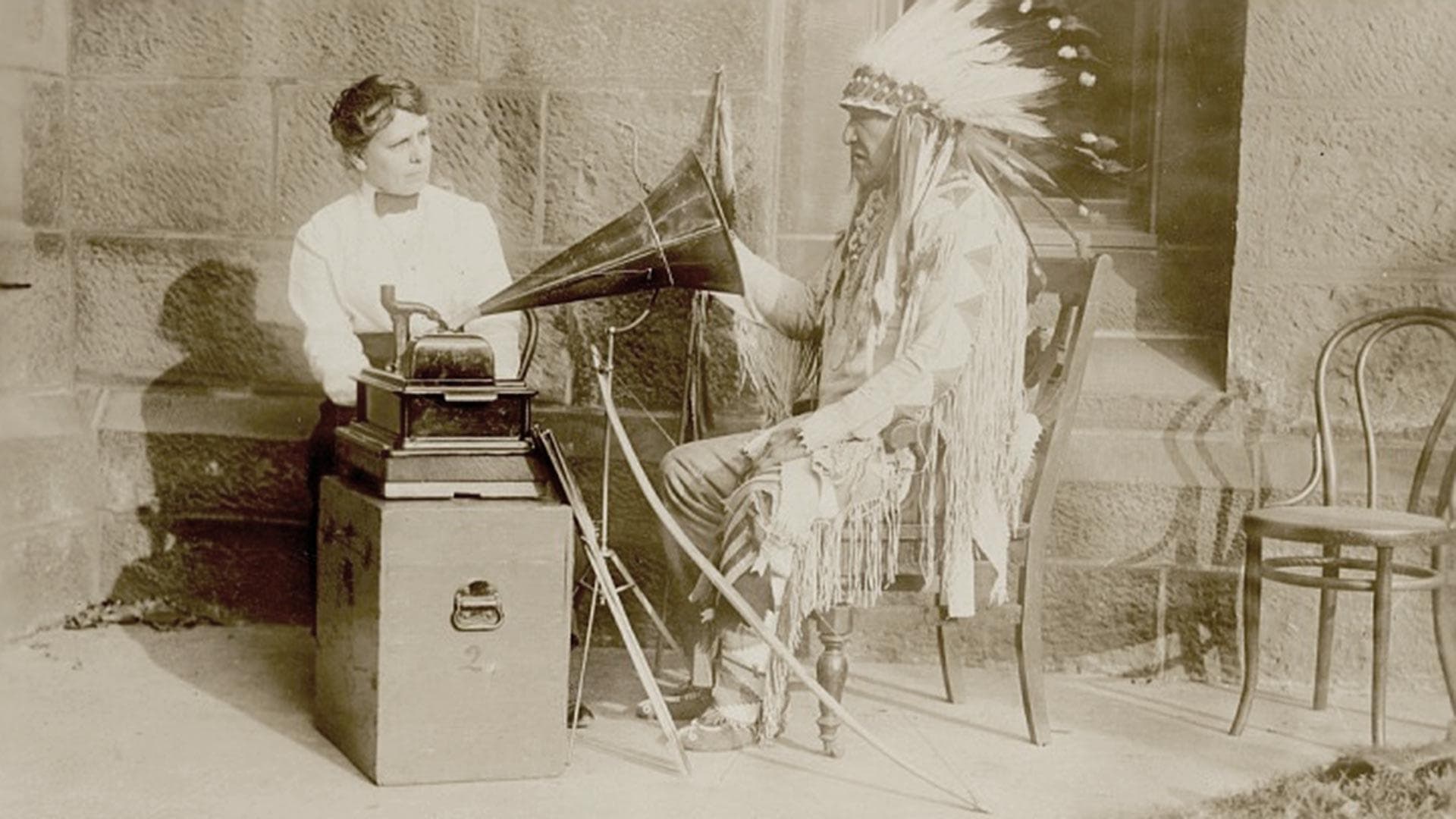 chief in Native dress with bow, arrows and lance listens to song being played on phonograph