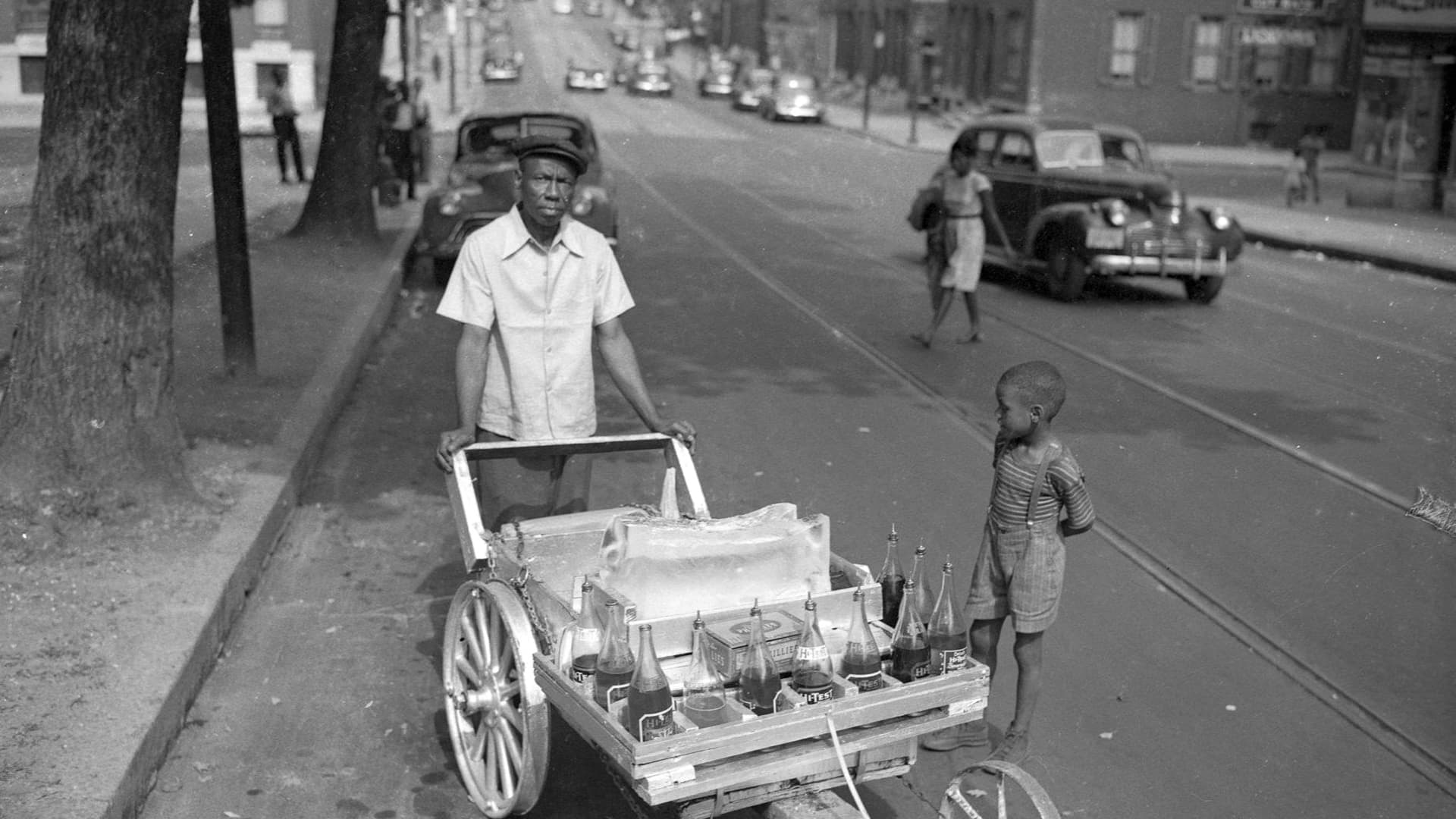 A young boy hovers expectantly near a snowball vendor in 1940s Harlem Park