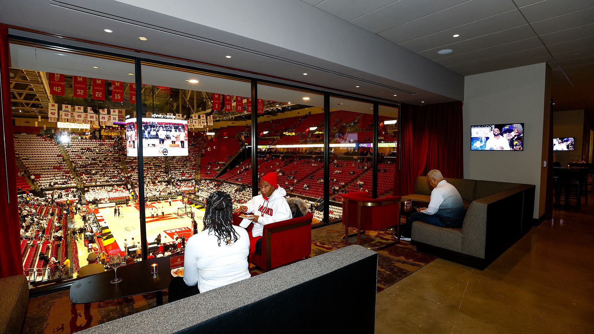 three fans sit in Maryland Club near windows overlooking basketball court