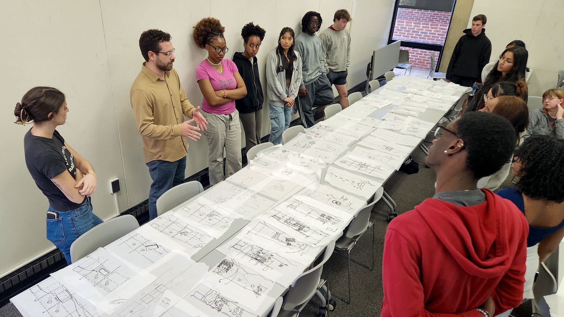 Students stand around a long rectangular desk and examine drawings