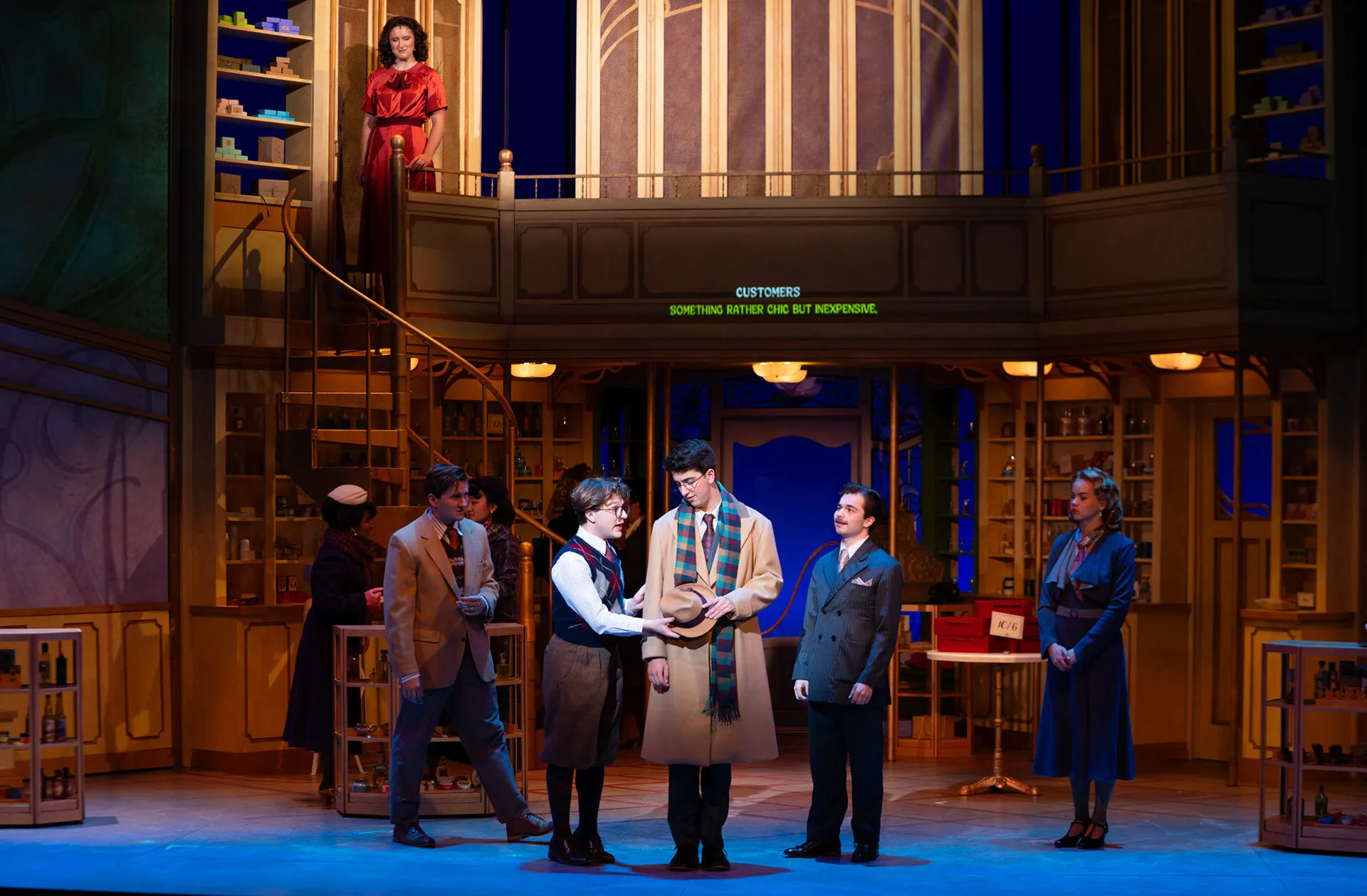 A woman in a red dress waits to go down the stairs while men in suits talk below; all part of a stage performance.