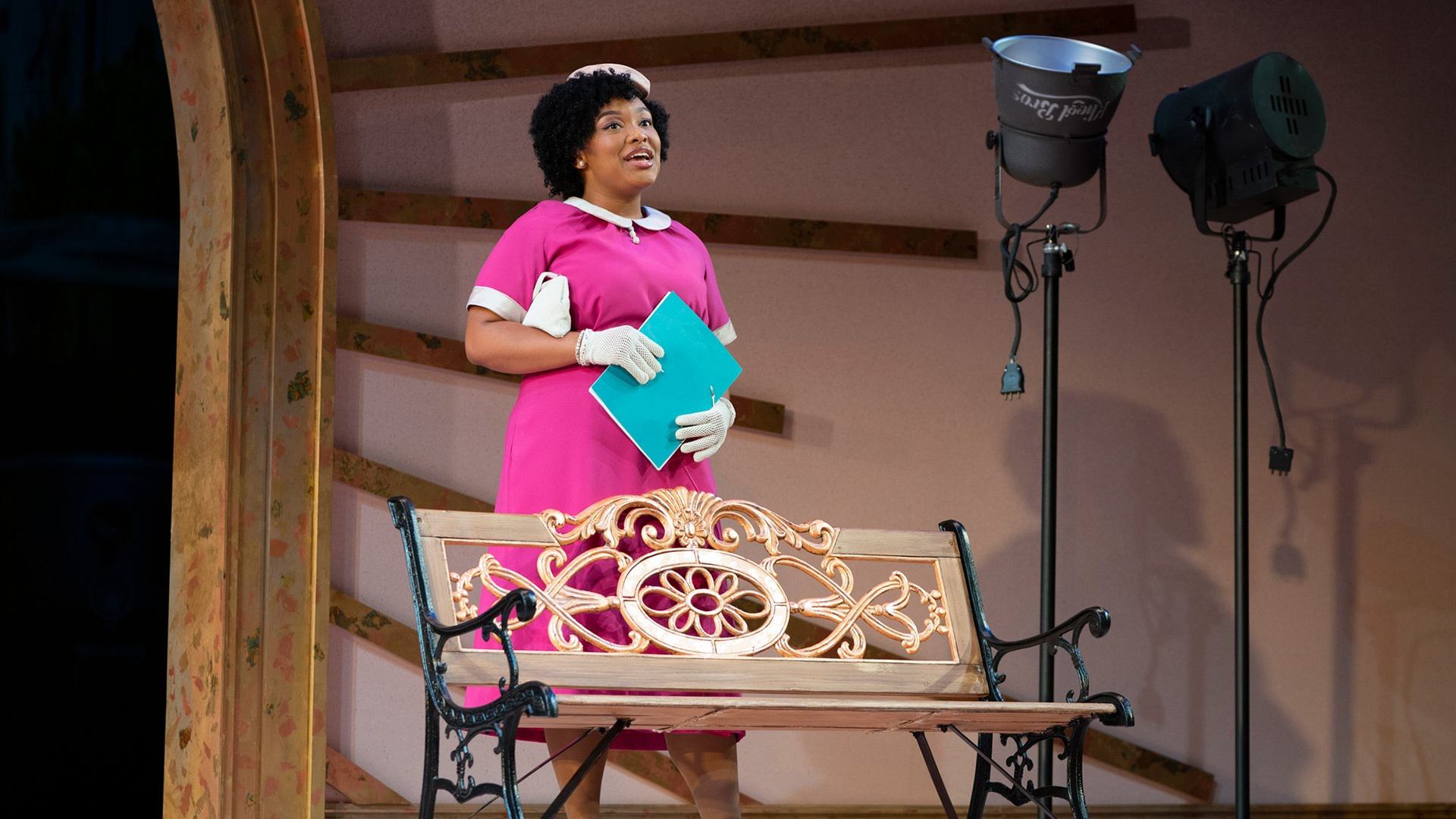 A Black woman holds a blue folder and stands behind a bench
