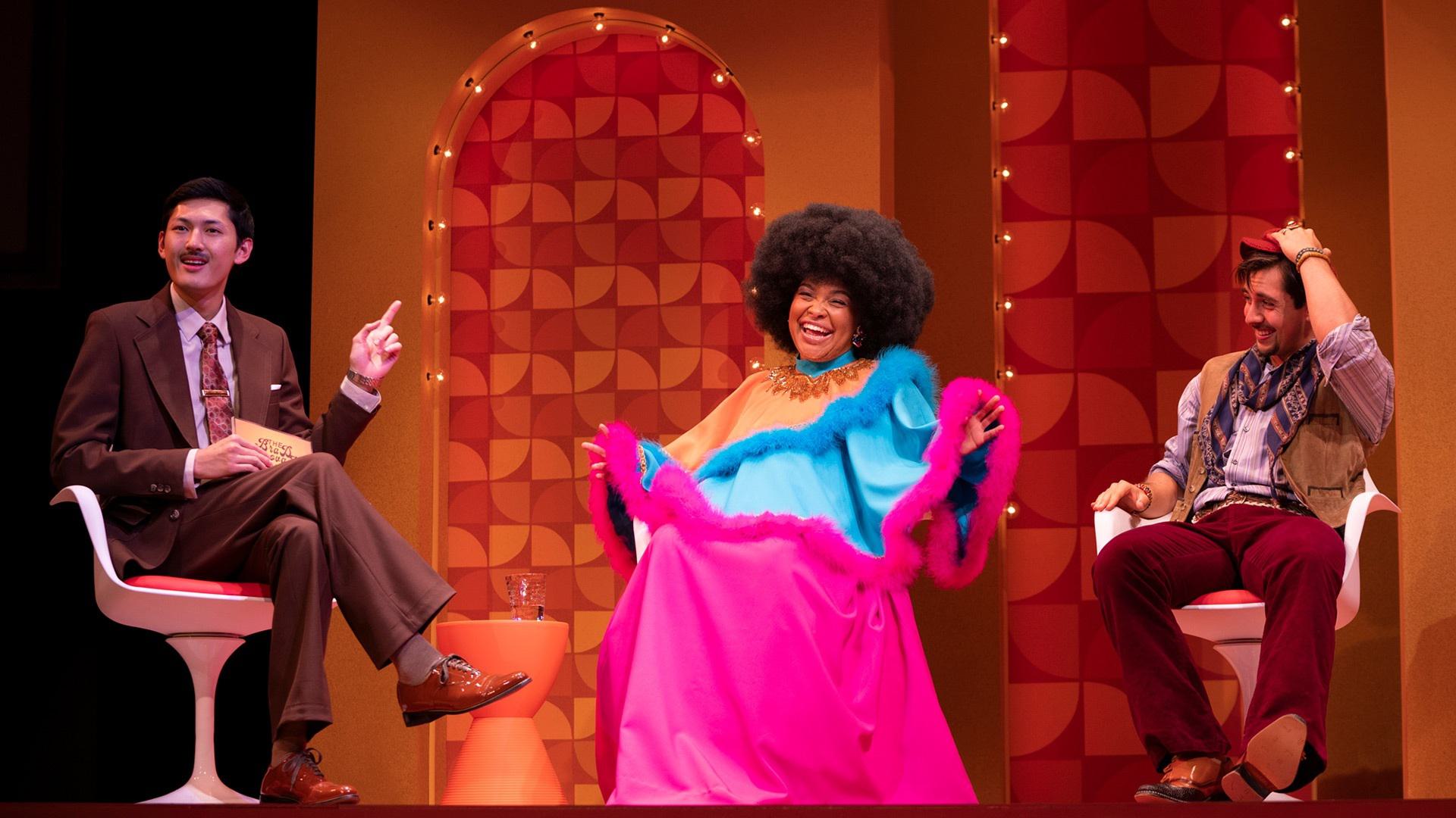 A woman in an Afro and bright pink fur-trimmed dress sits between two men