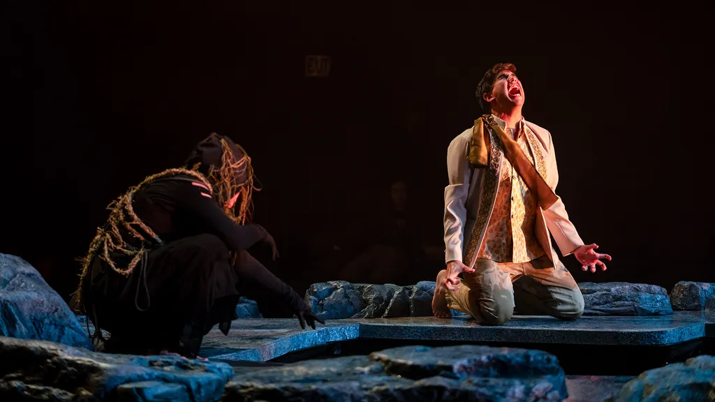 one actor kneels and yells while another crouches near pond on stage