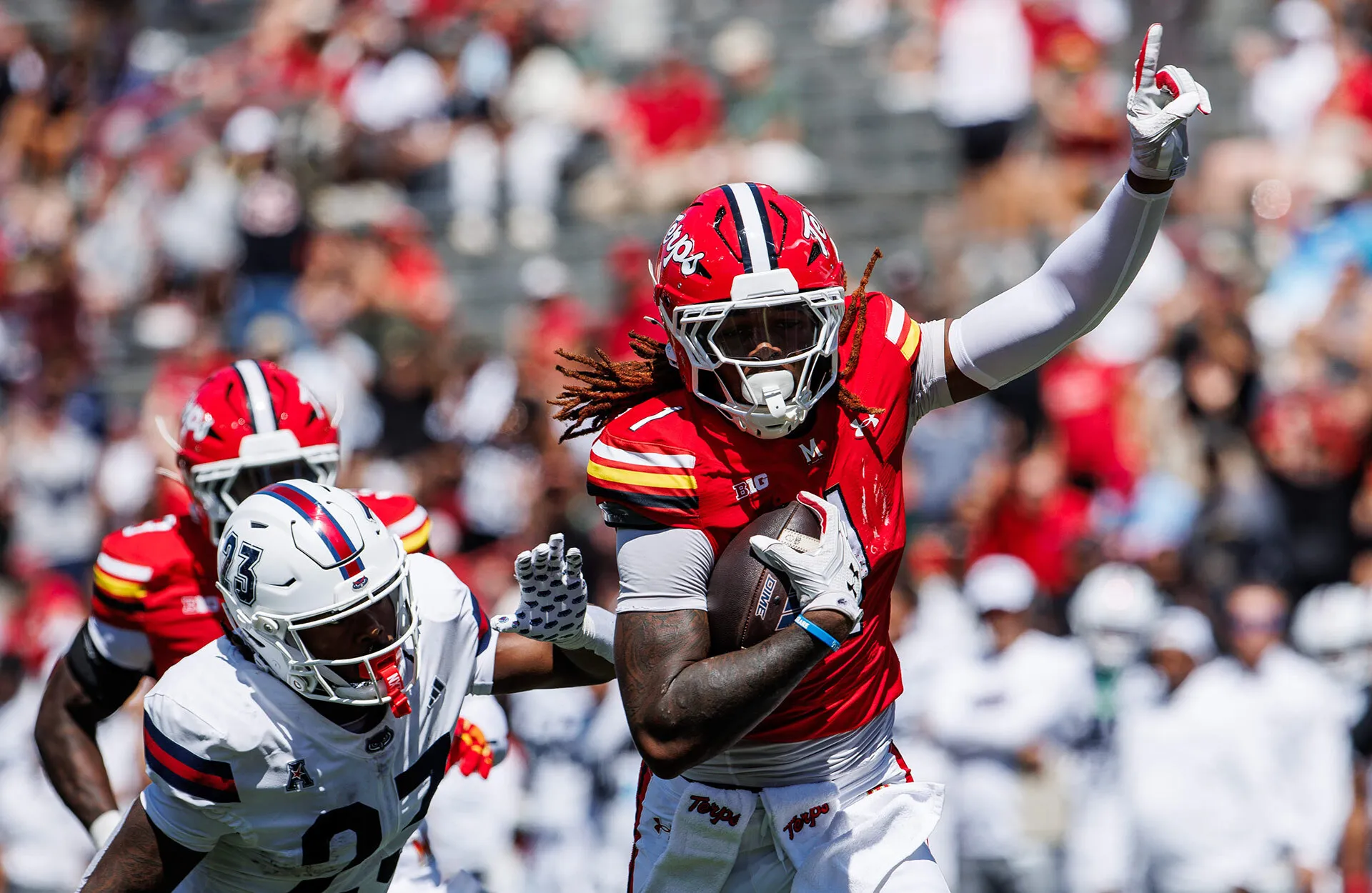 A Maryalnd football player runs with the ball while pointing to the sky and evading a tackler.