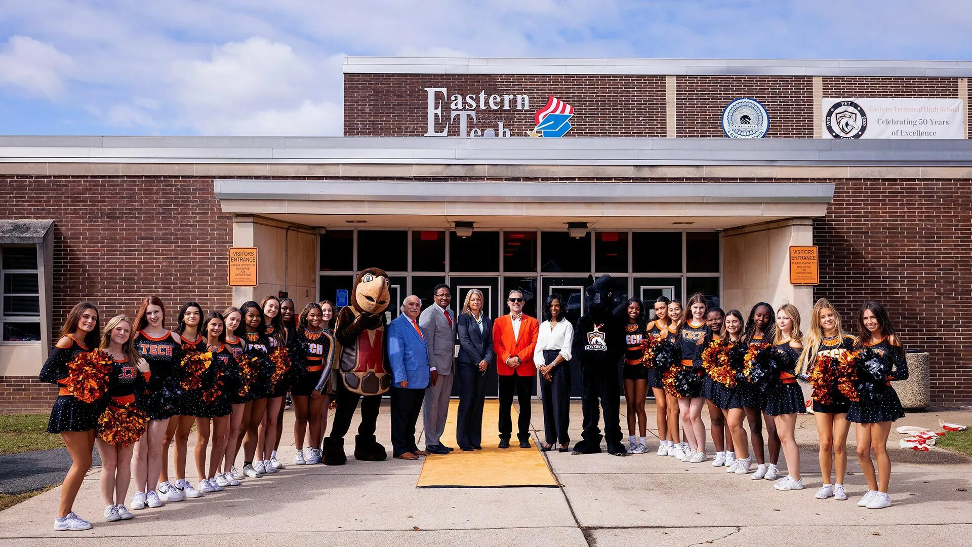 Cheerleaders, school officials and a mascot stand in front of a high school.