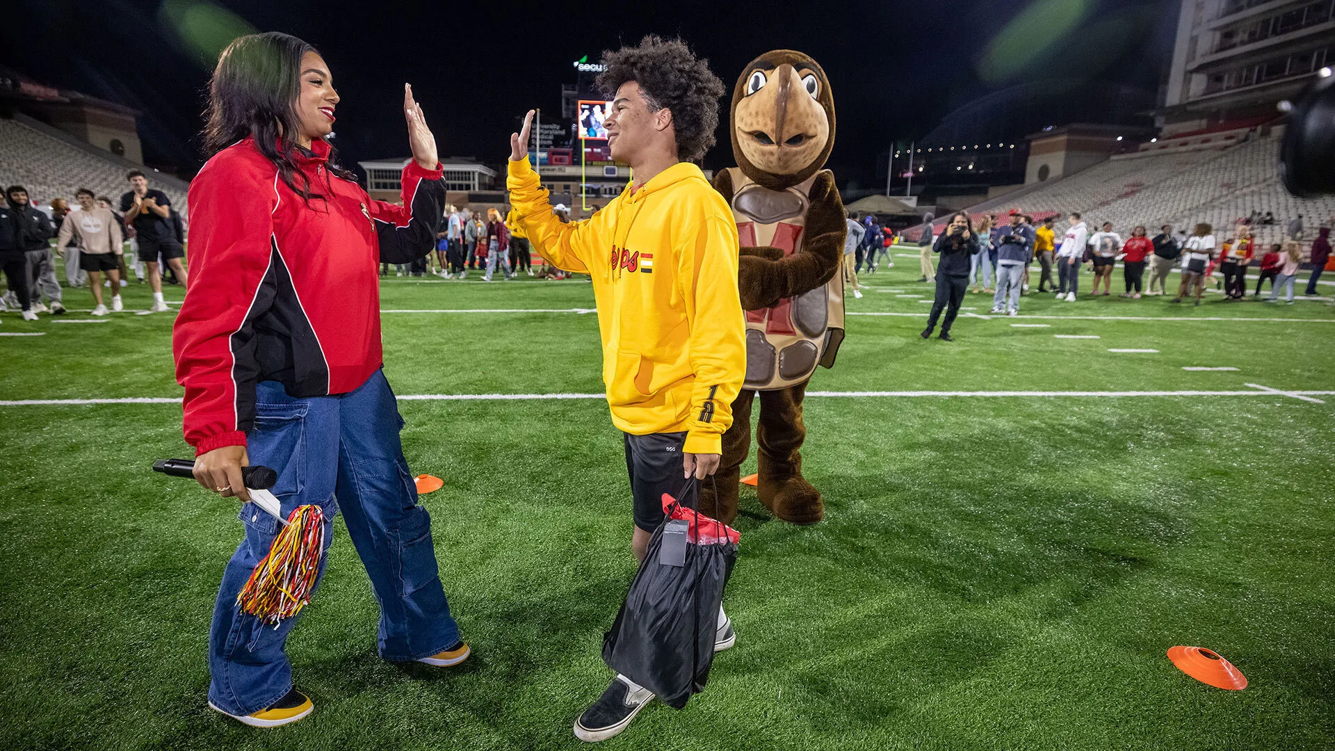 two people high five while a person dressed in a turtle mascot stands nearby on a football field
