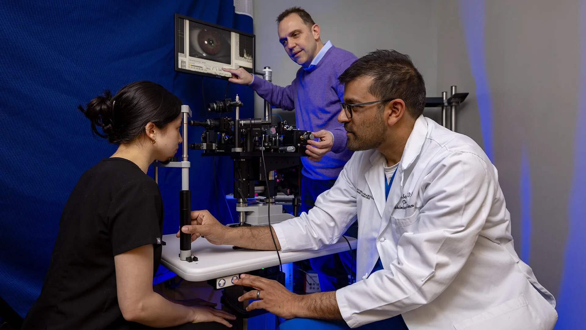 Dr. Osamah J. Saeedi wears a white coat and works with a patient on an eye-screening machine