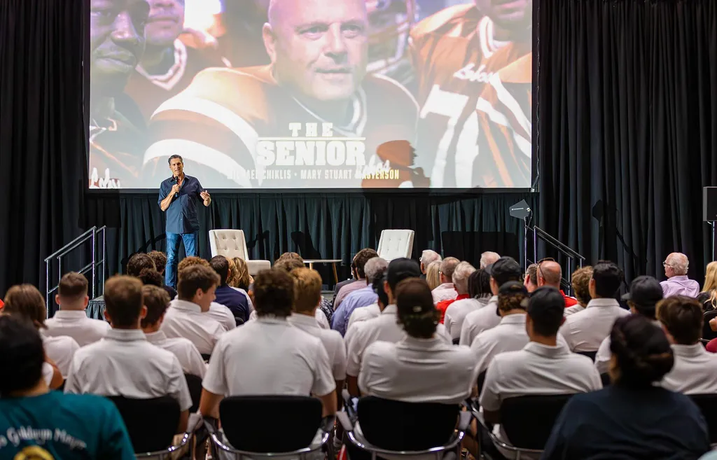 Man talks to audience while standing in front of movie screen showing a football player