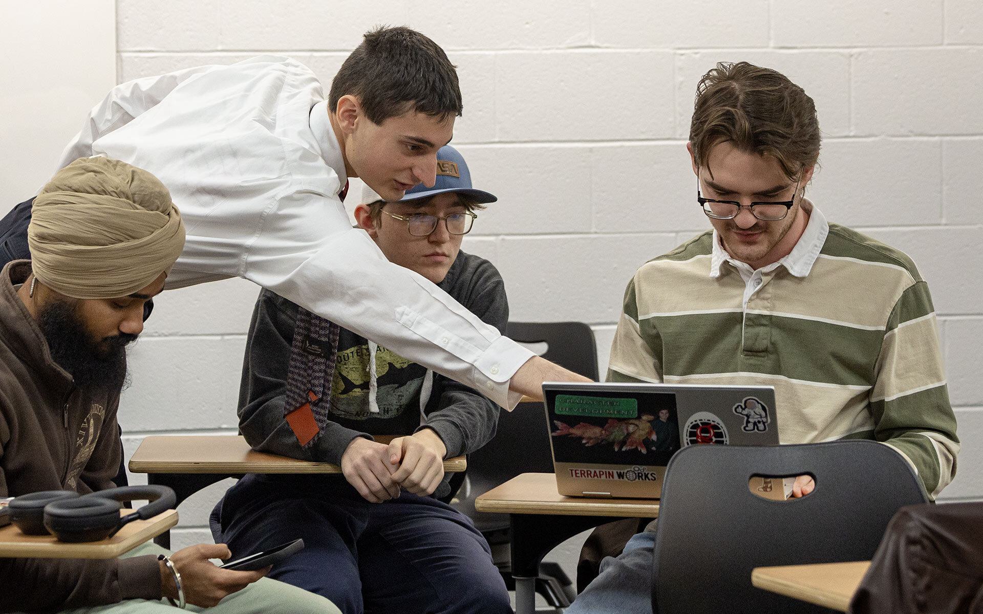 students sit at desks, with one student pointing to laptop