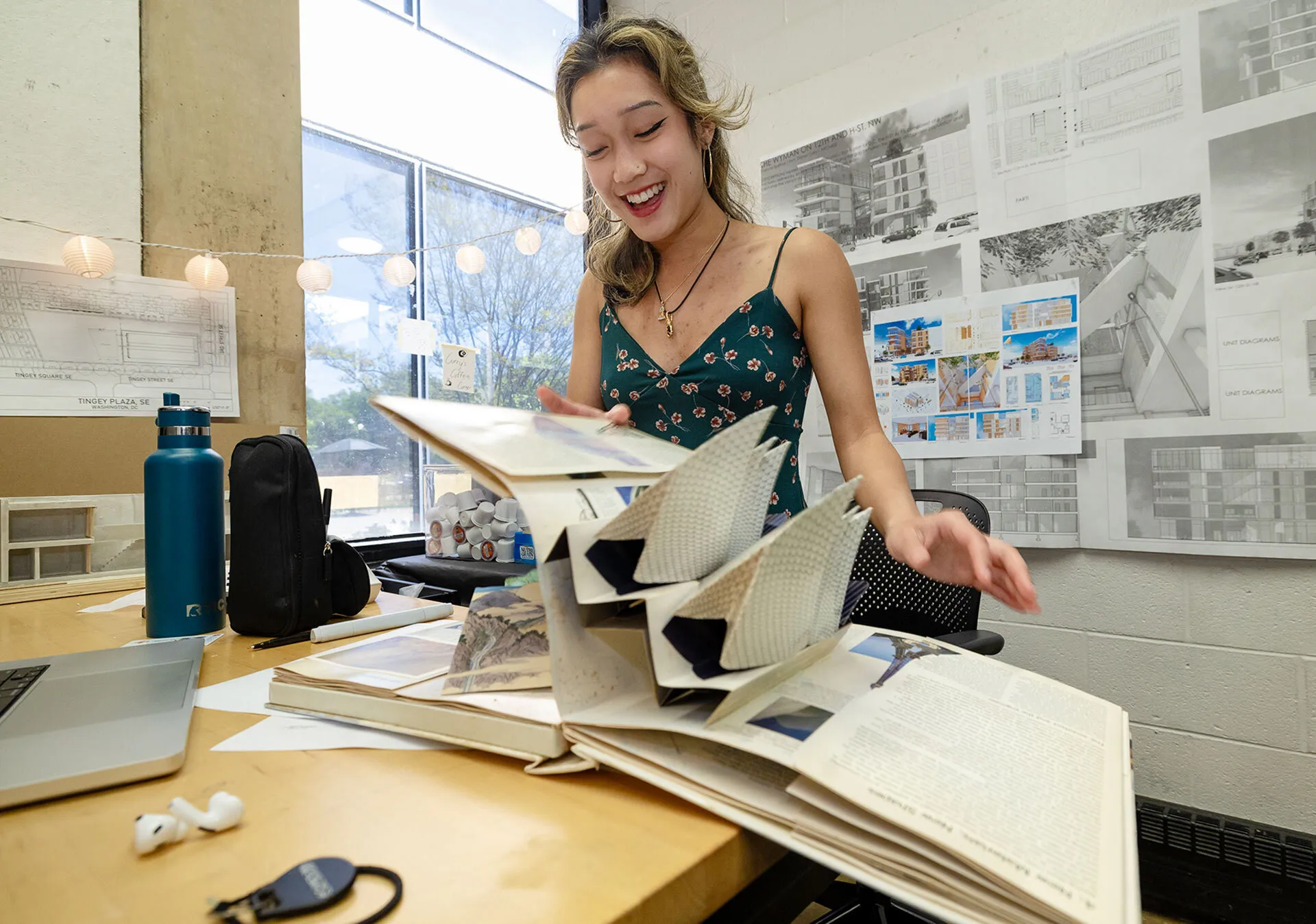 Evelyne Sokhon flips through an architecture book at her desk with water bottles and architectural drawings in the background