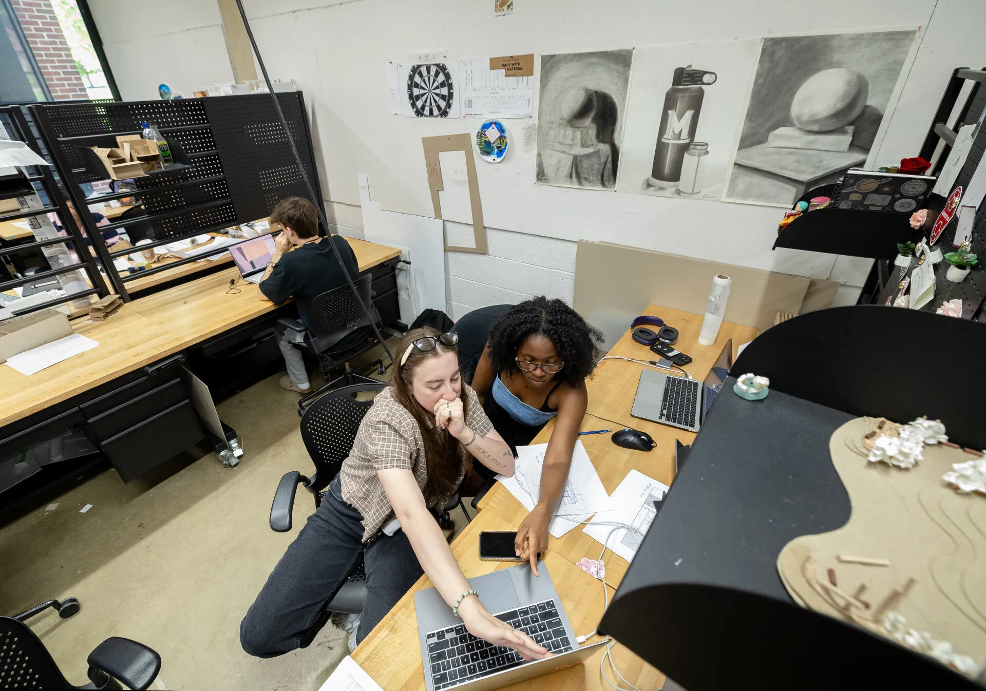 Overhead shot of two students pointing at a laptop on a desk with black and white still life drawings on the wall behind them