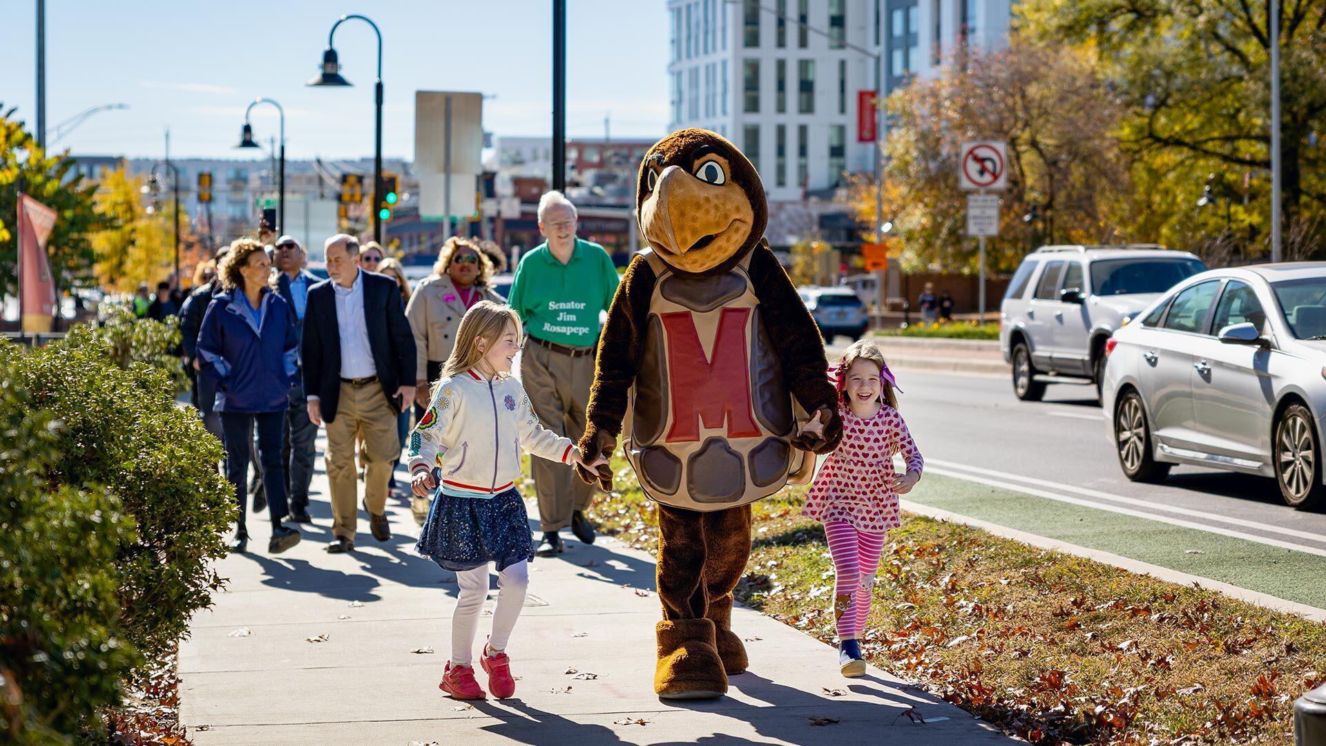 two girls hold hands with Testudo, leading crowd down Baltimore Avenue