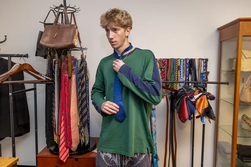 A student in a green long-sleeved shirt tries on a blue tie with racks of ties and purses behind him