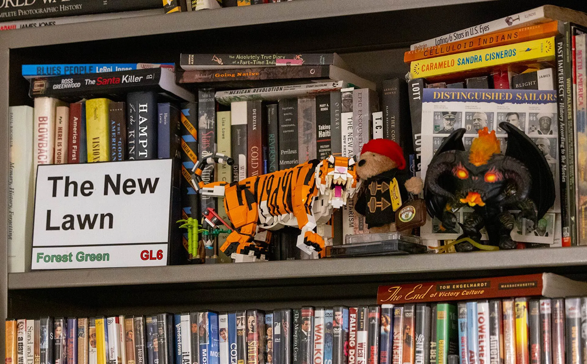 A shelf with books and DVDs with a sign saying "The New Lawn", a Lego tiger, Paddington the bear and a Balrog