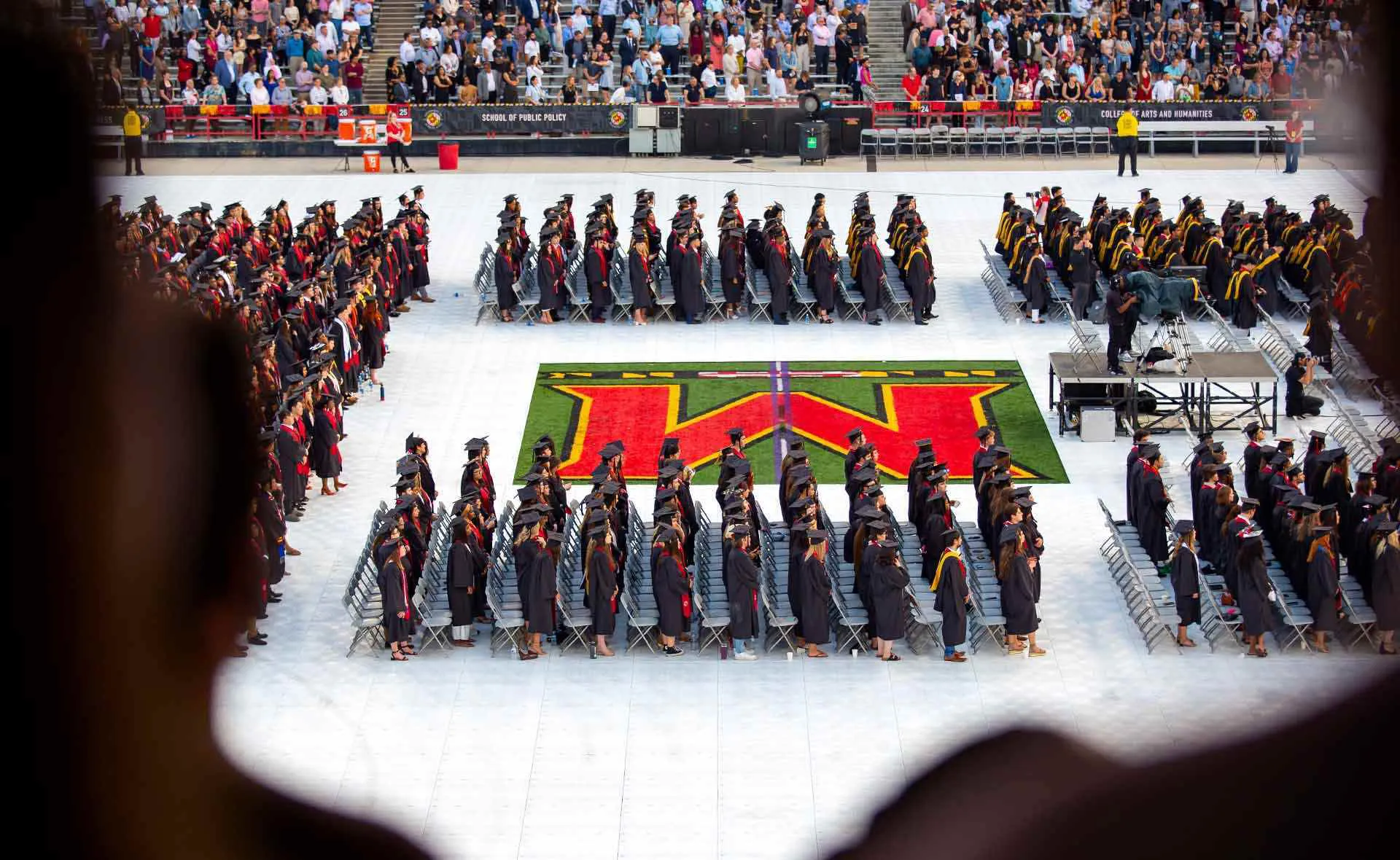 students in grad regalia wait for ceremony on SECU Stadium field