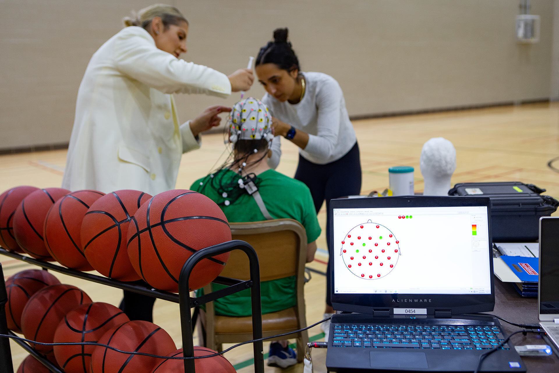 two researchers look at EEG cap on man's head, with basketballs and laptop in foreground