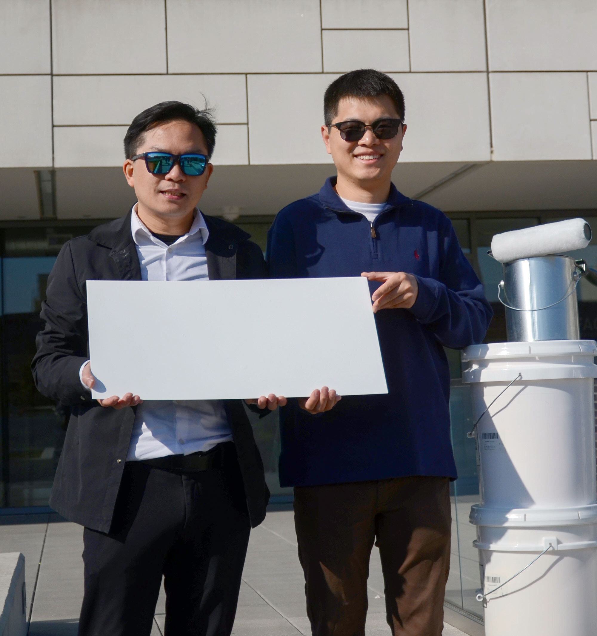 two men hold a white rectangular piece of metal in front of a white building