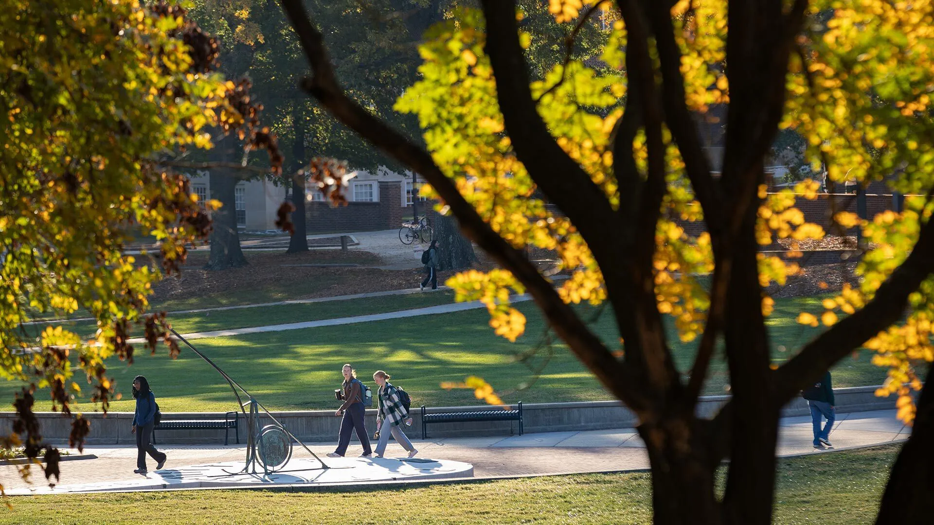 students walk past sundial with yellow leaves on trees