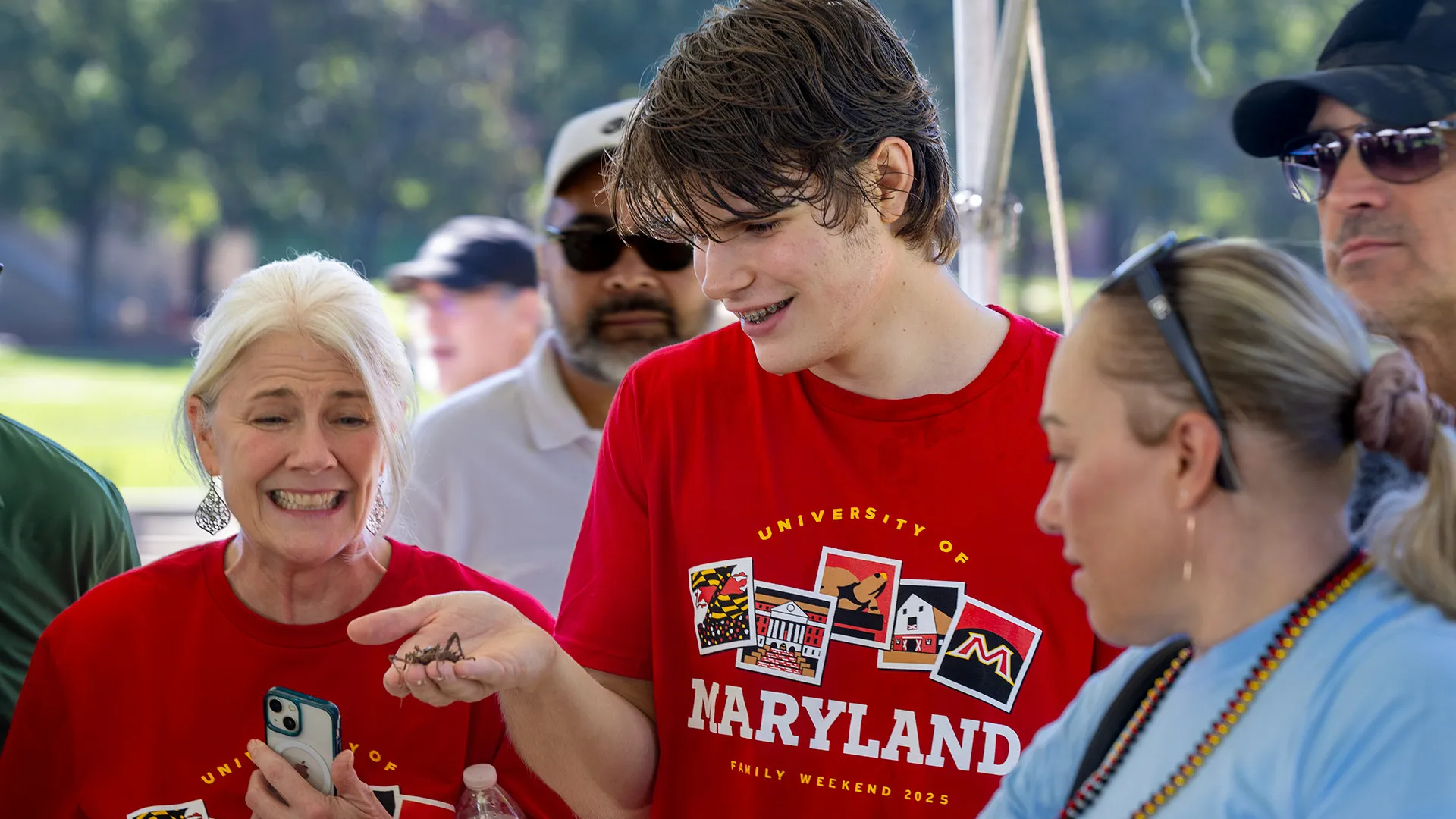 A young man holds a large insect while a woman winces.