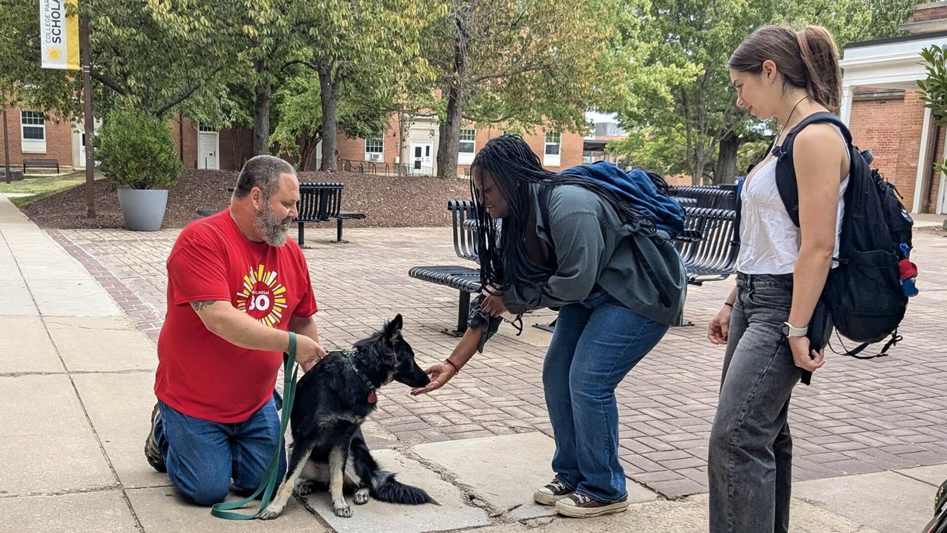 man in red shirt holds German shepherd on leash as one woman holds her hand out to the dog and another looks on