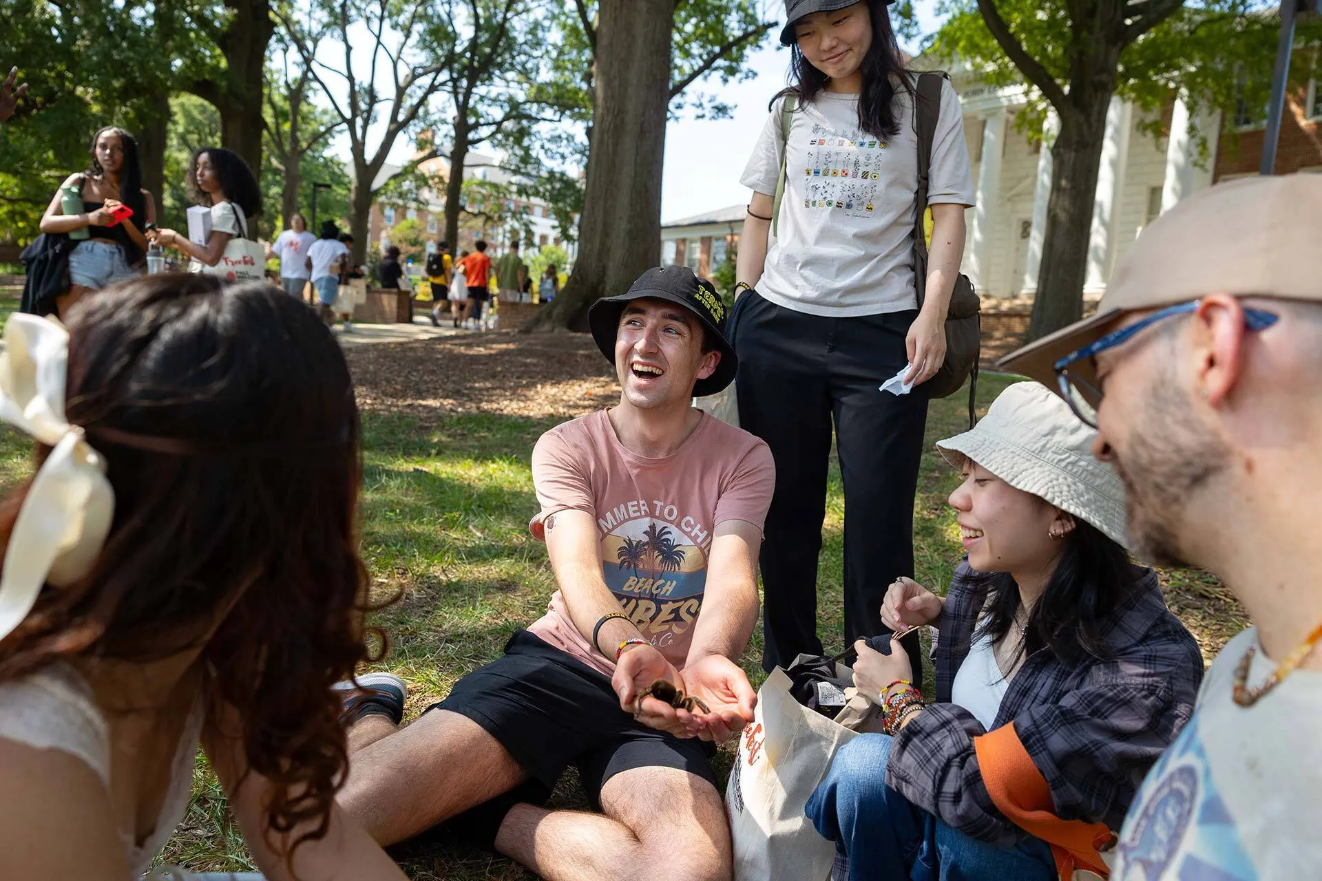 young man in black bucket hat and pink shirt smiles as he holds giant spider and other students observe mirthfully