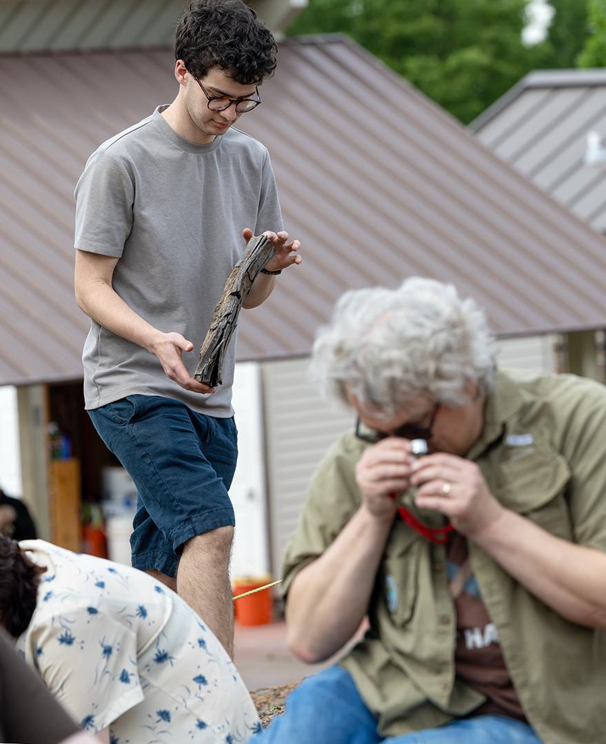 A student holds a piece of fossilized wood while a professor uses a tool to examine a piece of rock