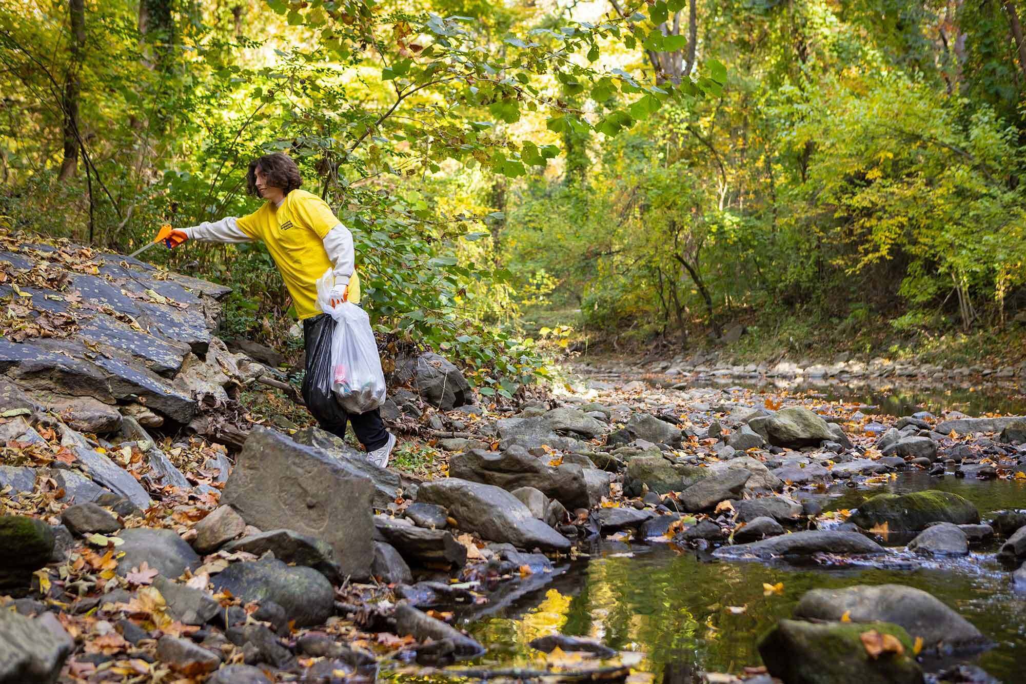 man wearing yellow shirt picks up trash in nature near a stream