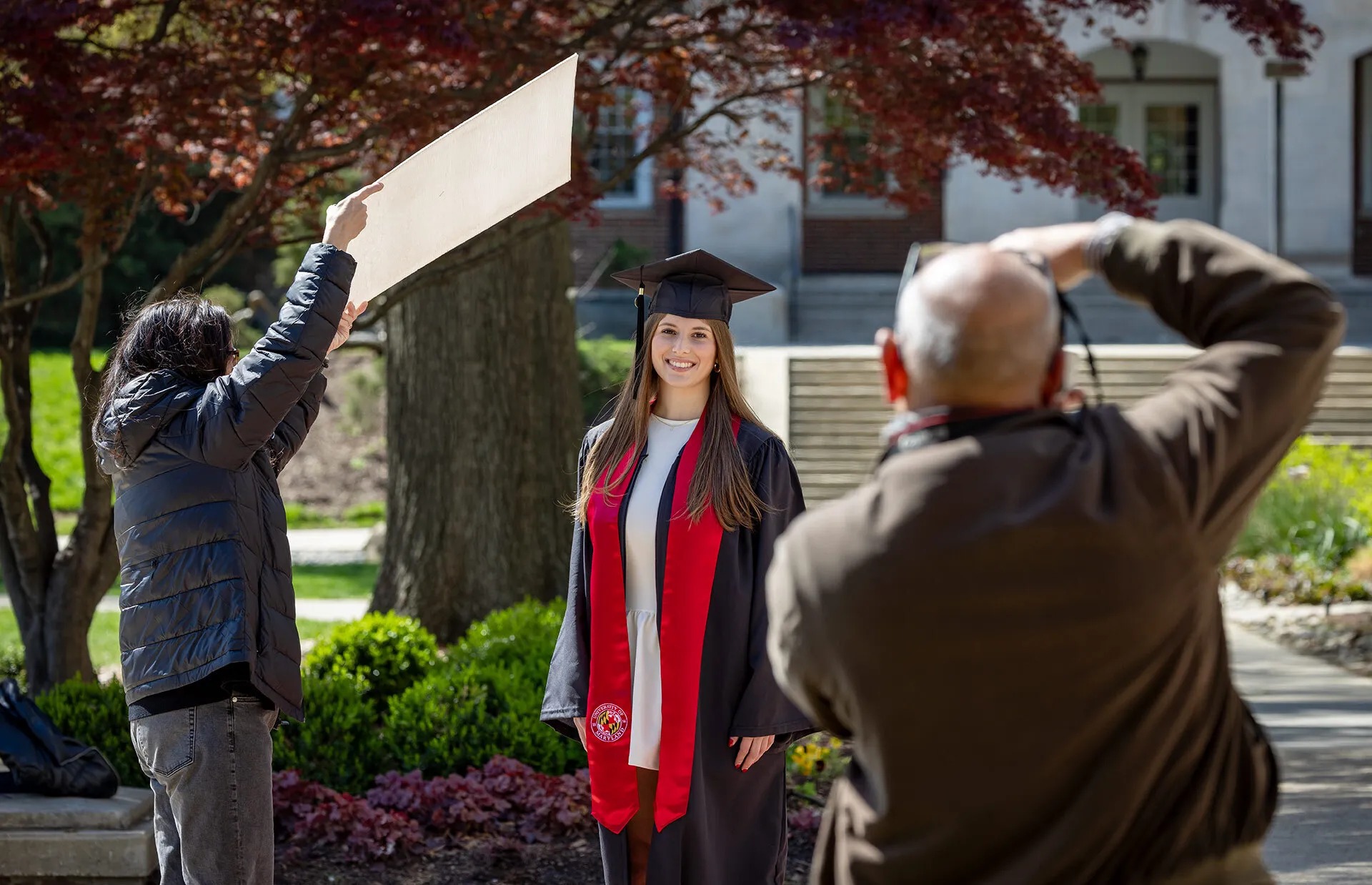 A person holds a white posterboard to shield the sun over a student in a cap and gown as a photographer takes a photo