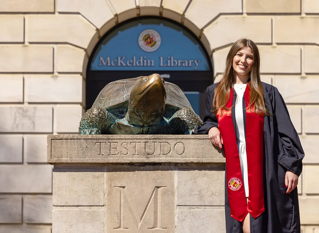 A student in a graduation gown poses next to the bronze Testudo statue