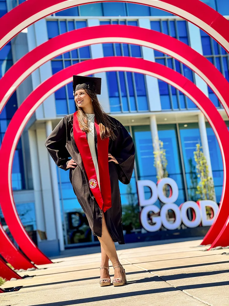 A student in graduation cap and gown stands inside red rings with Do Good in the background