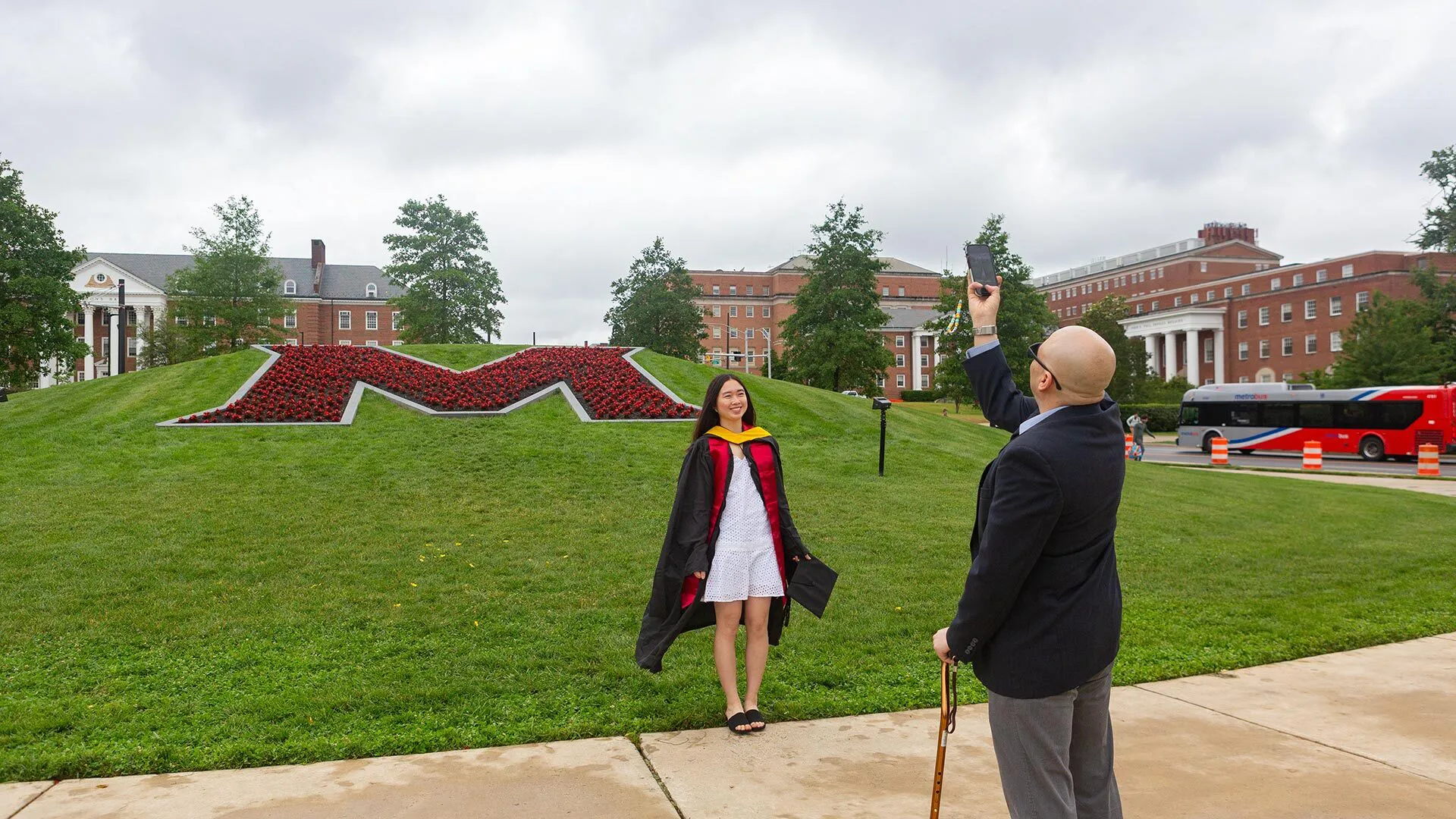 man takes a photo of a student wearing grad regalia in front of the red flower M