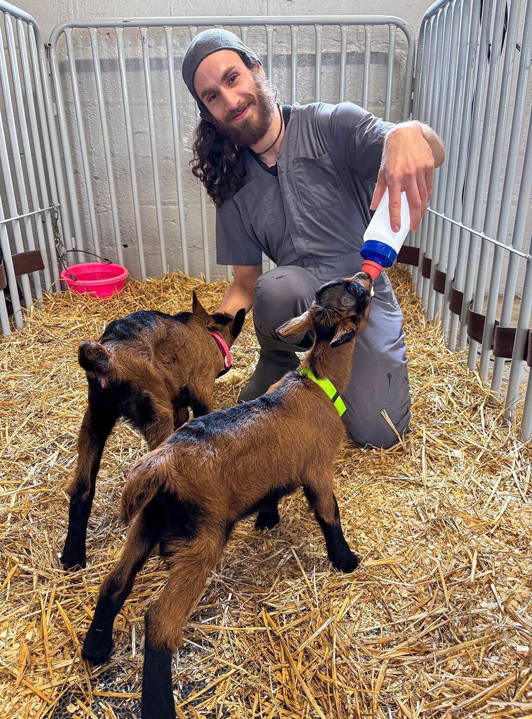 Gregory Garin feeds a bottle to goat kids while wearing a gray jumpsuit and standing on hay