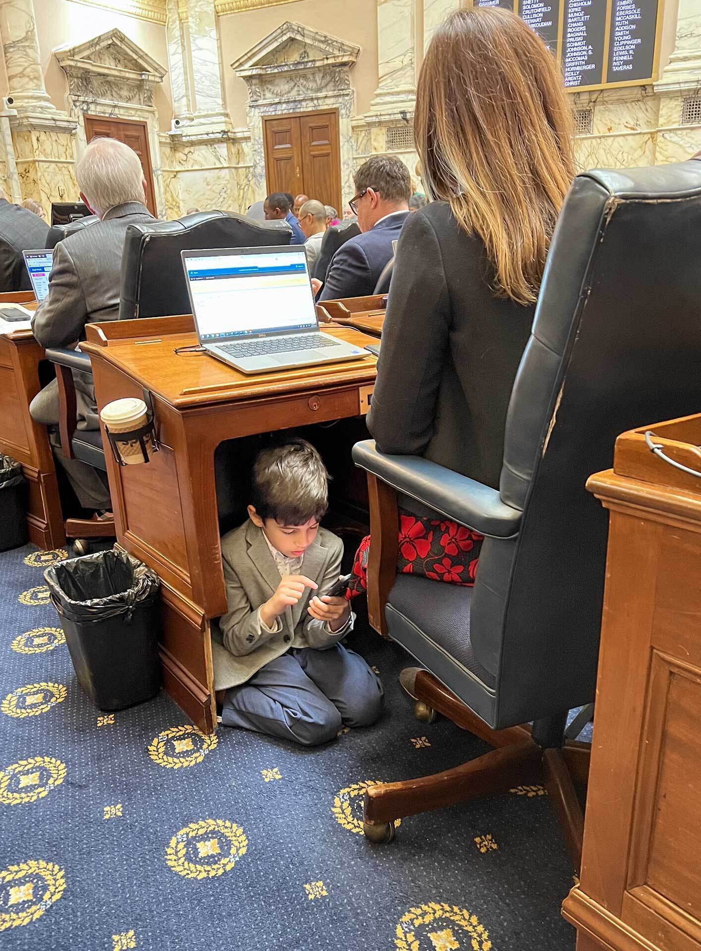 young boy looks at phone under desk as woman works on laptop